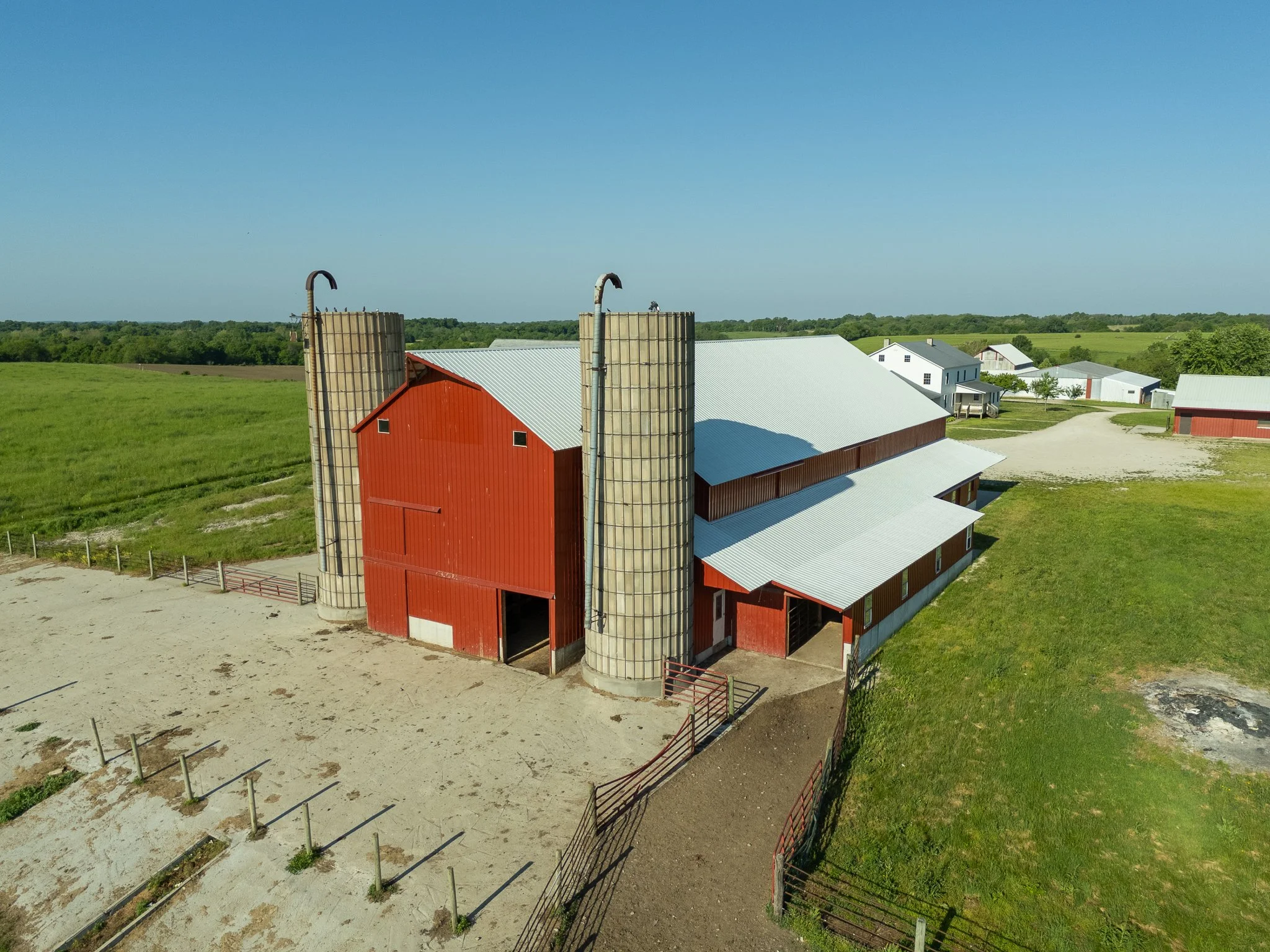 Aerial view of a red barn with two silos in a rural farm setting, surrounded by green fields and other farm buildings under a clear blue sky.