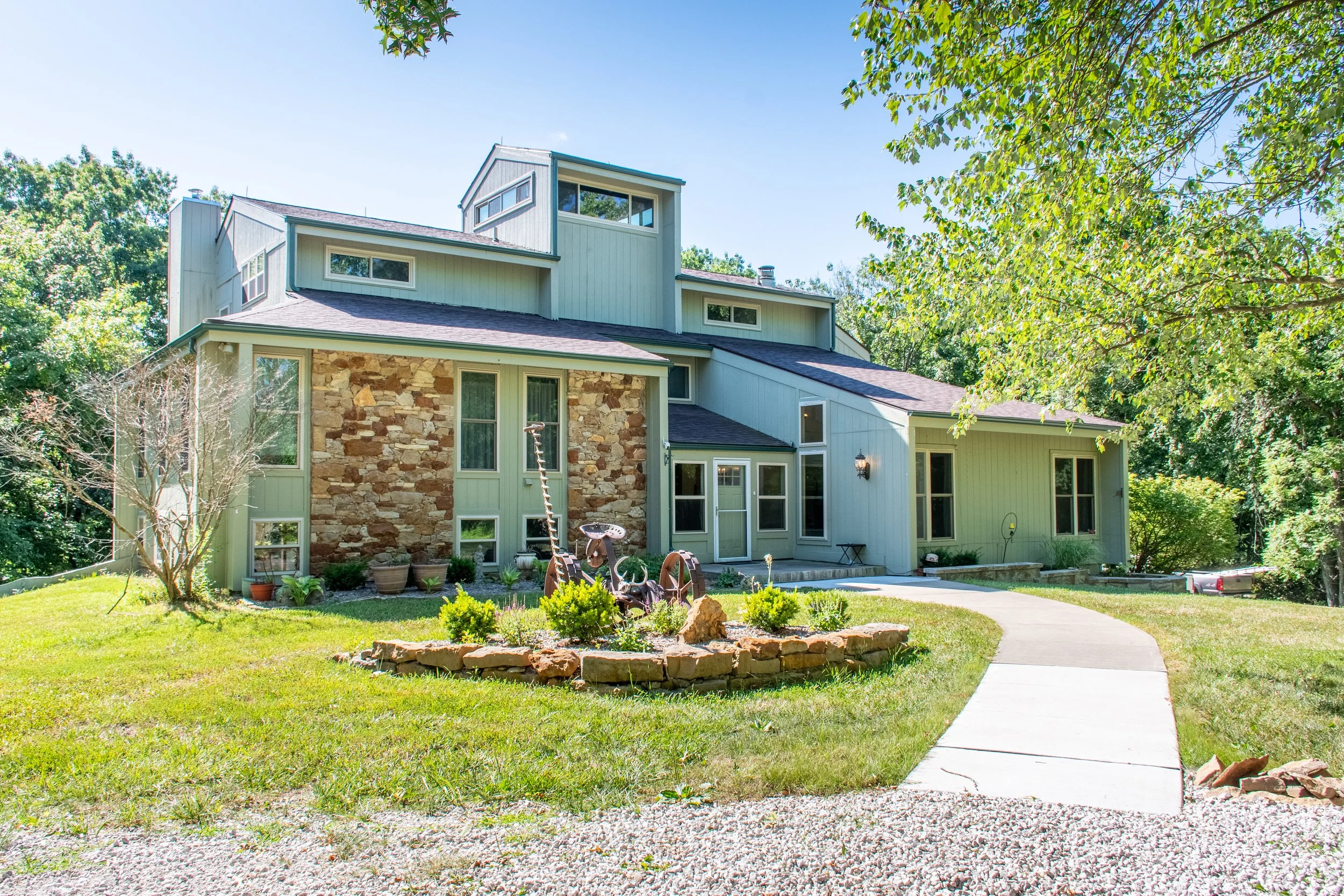 A large modern house with stone and light-colored siding, surrounded by a grassy yard and trees, with a concrete walkway leading to the front door.