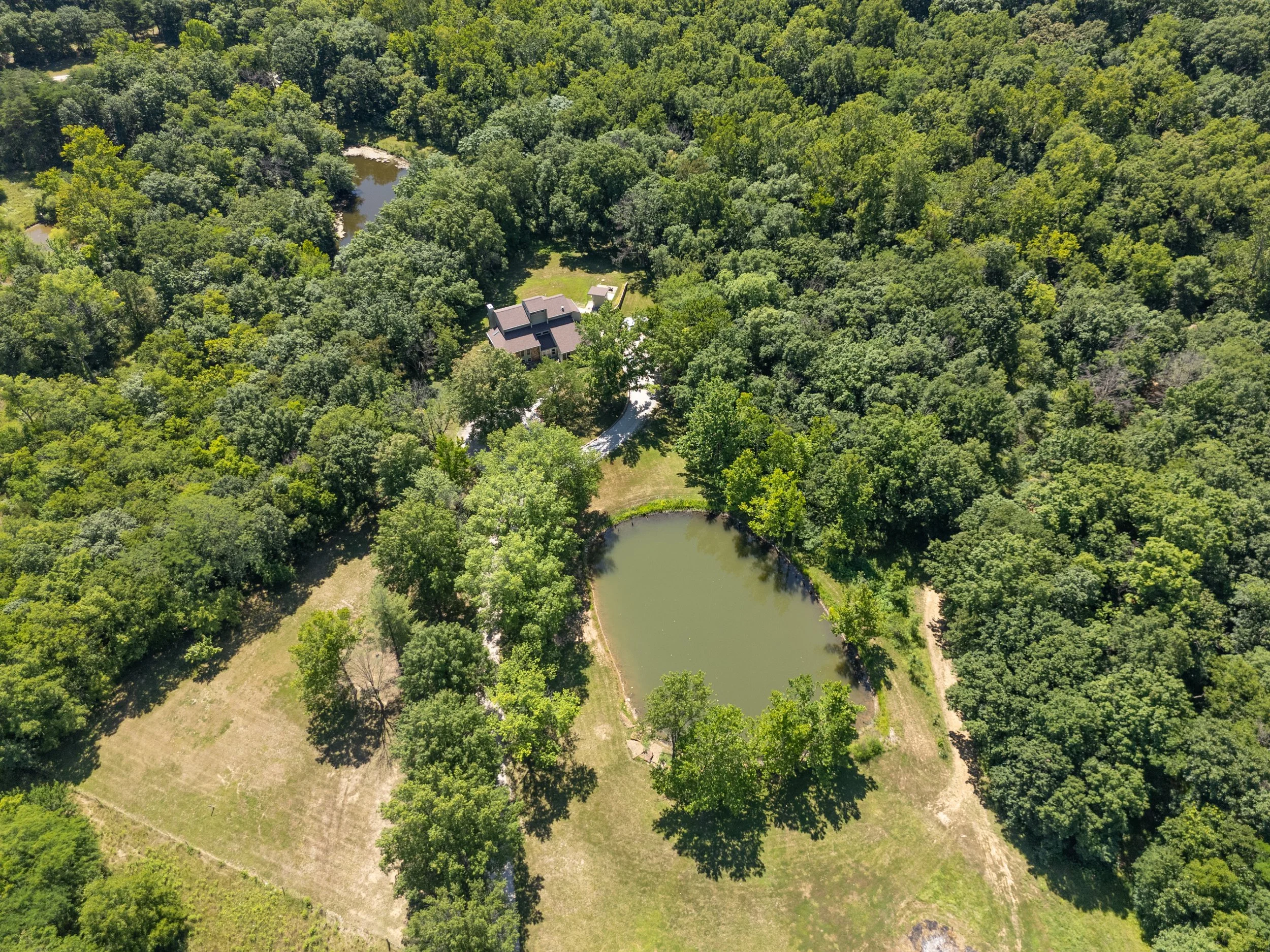 Aerial view of a house surrounded by dense green trees, with a small pond and a grassy area nearby.
