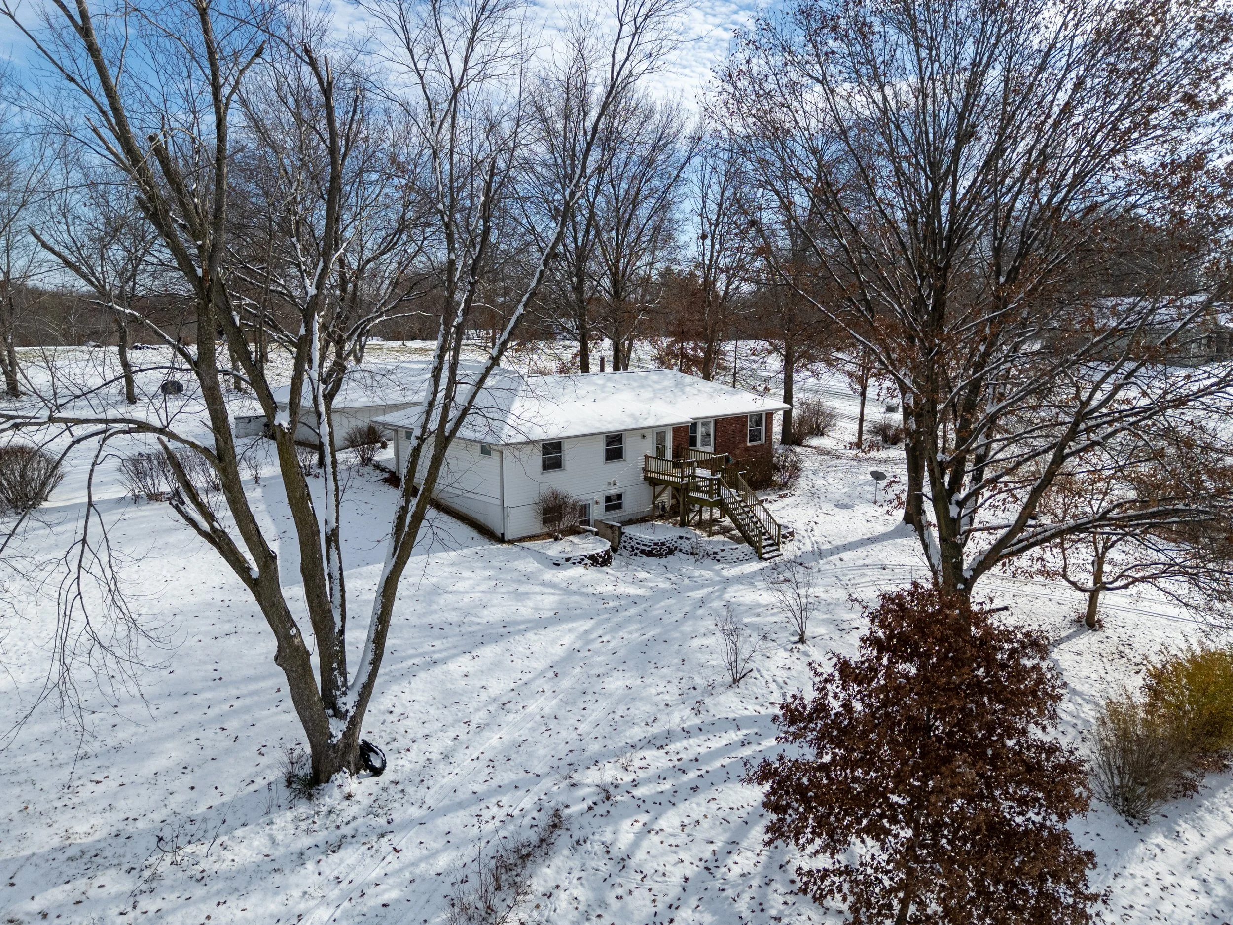 A snow-covered backyard with large trees, a white house with a brick section, and a deck with stairs, under a partly cloudy sky.