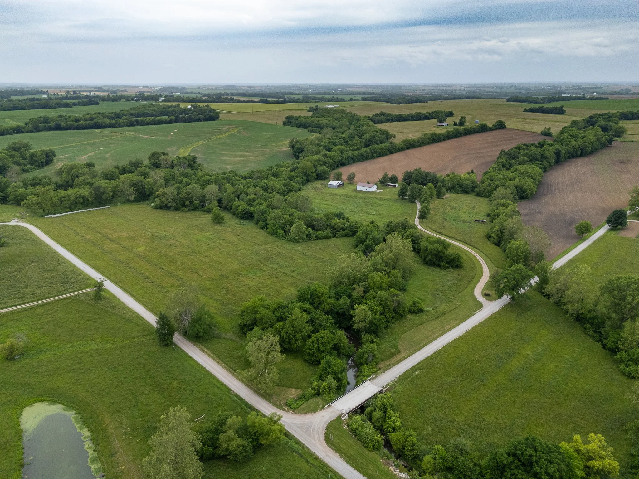 Aerial view of a rural landscape featuring winding roads, green fields, wooded areas, barns, and farmland with cloudy sky overhead.