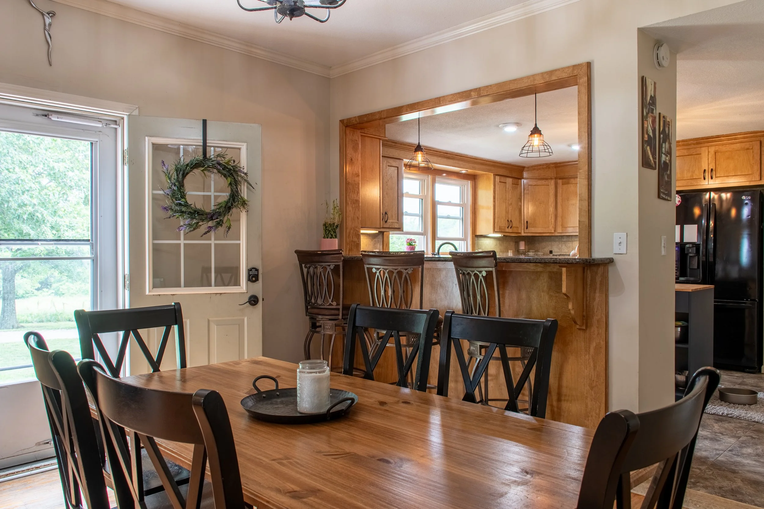 Kitchen and dining area with wooden table, black chairs, and a bar counter with stools, featuring warm lighting and natural light from windows.