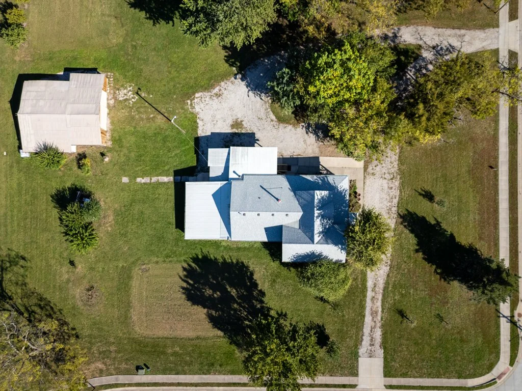 An aerial view of a residential property with a house, a detached shed, trees, and a lawn with a garden patch. There are sidewalks on the streets bordering the yard.