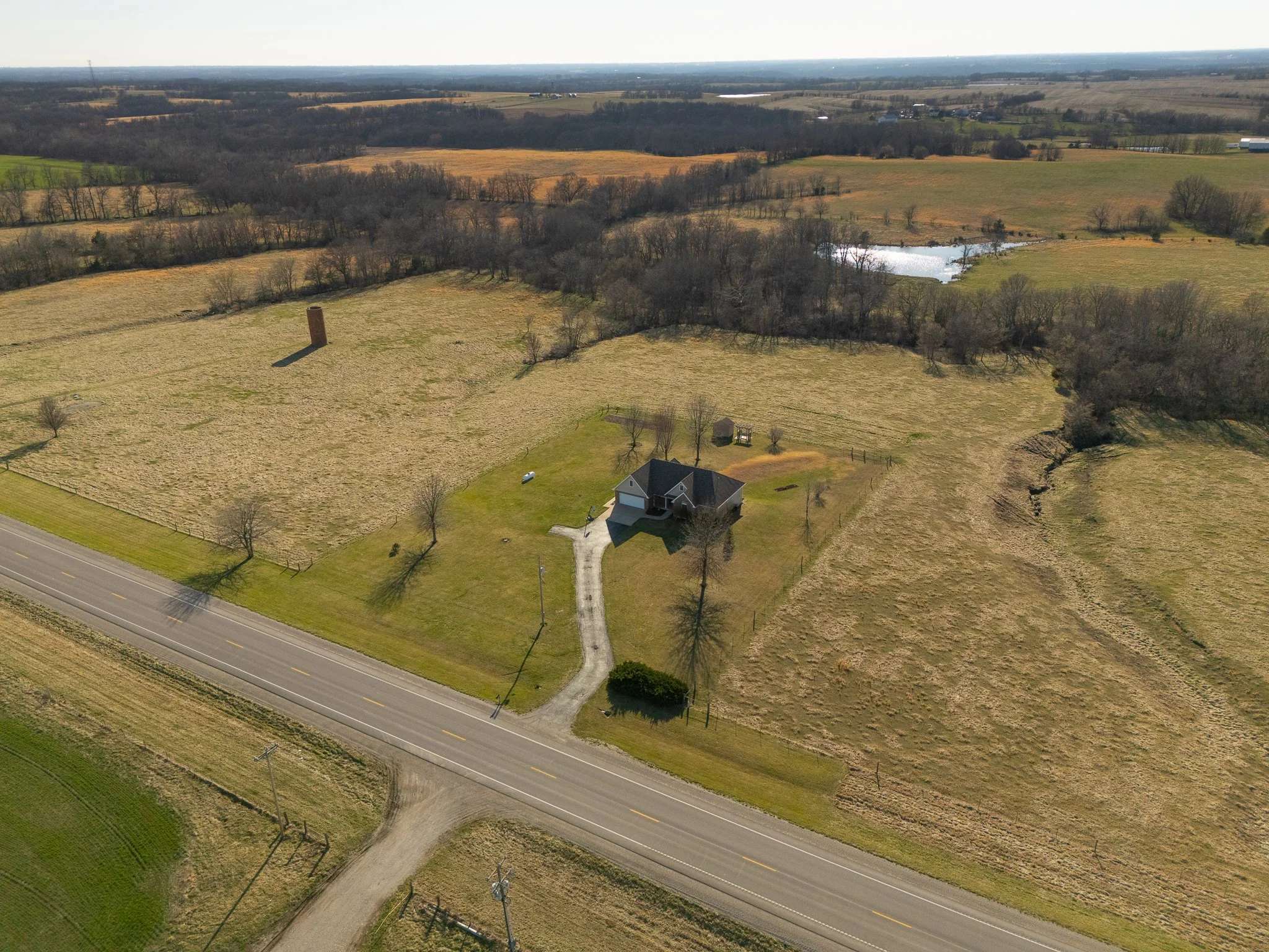 Aerial view of a rural landscape with a house, a driveway, open fields, a road, trees, and small bodies of water in the background.