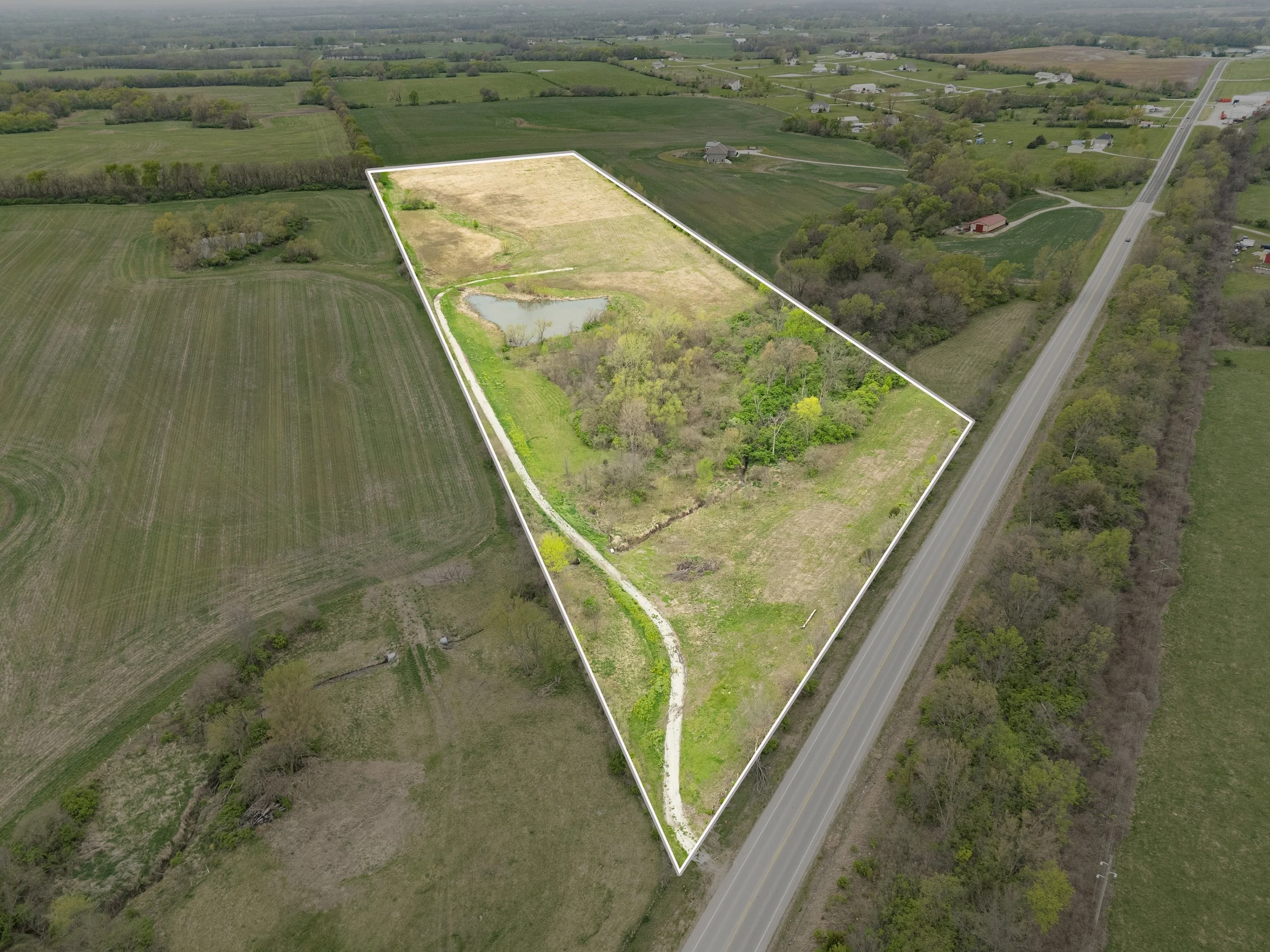 Aerial view of a large rural property outlined in white, with a pond, trees, open fields, and a gravel road winding through.