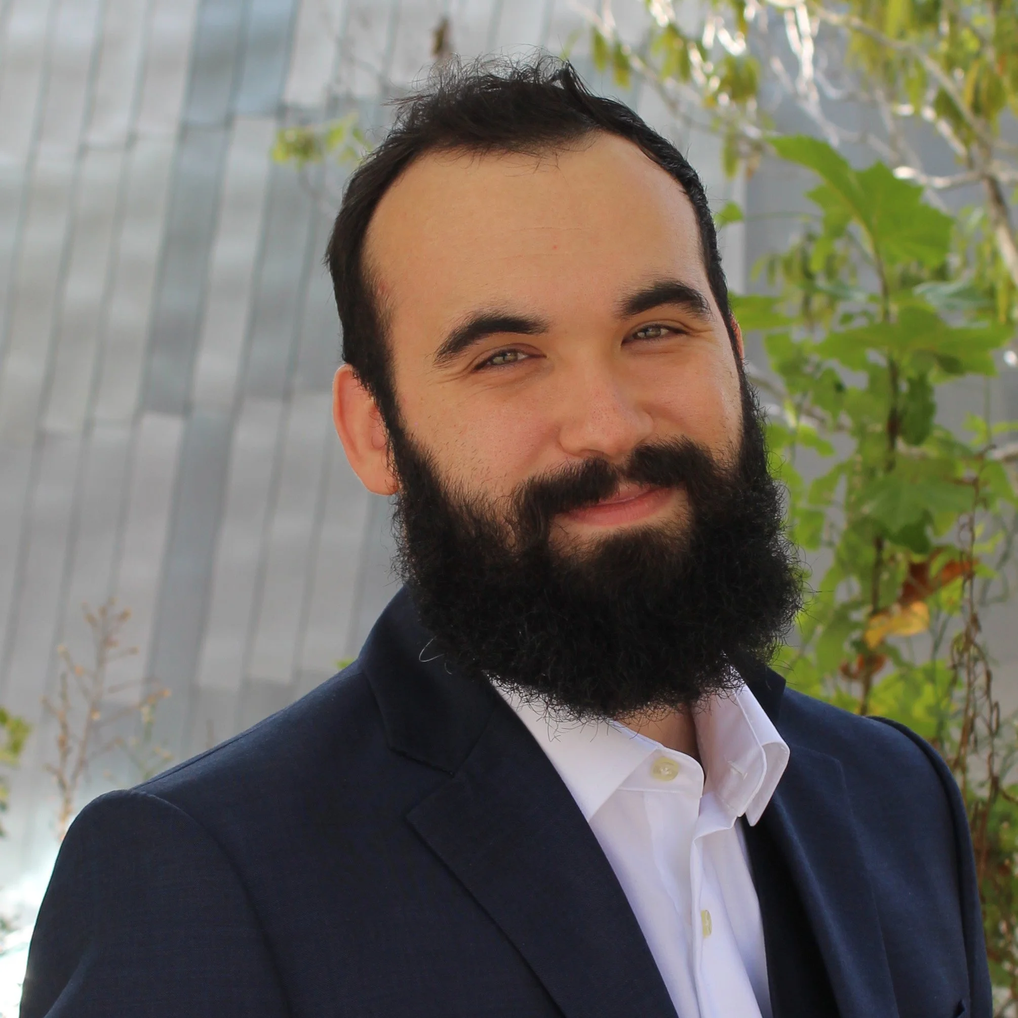 A Portrait of Raiden page, a man with dark hair and a full beard, wearing a navy blazer and white dress shirt, standing outdoors with green foliage in the background.