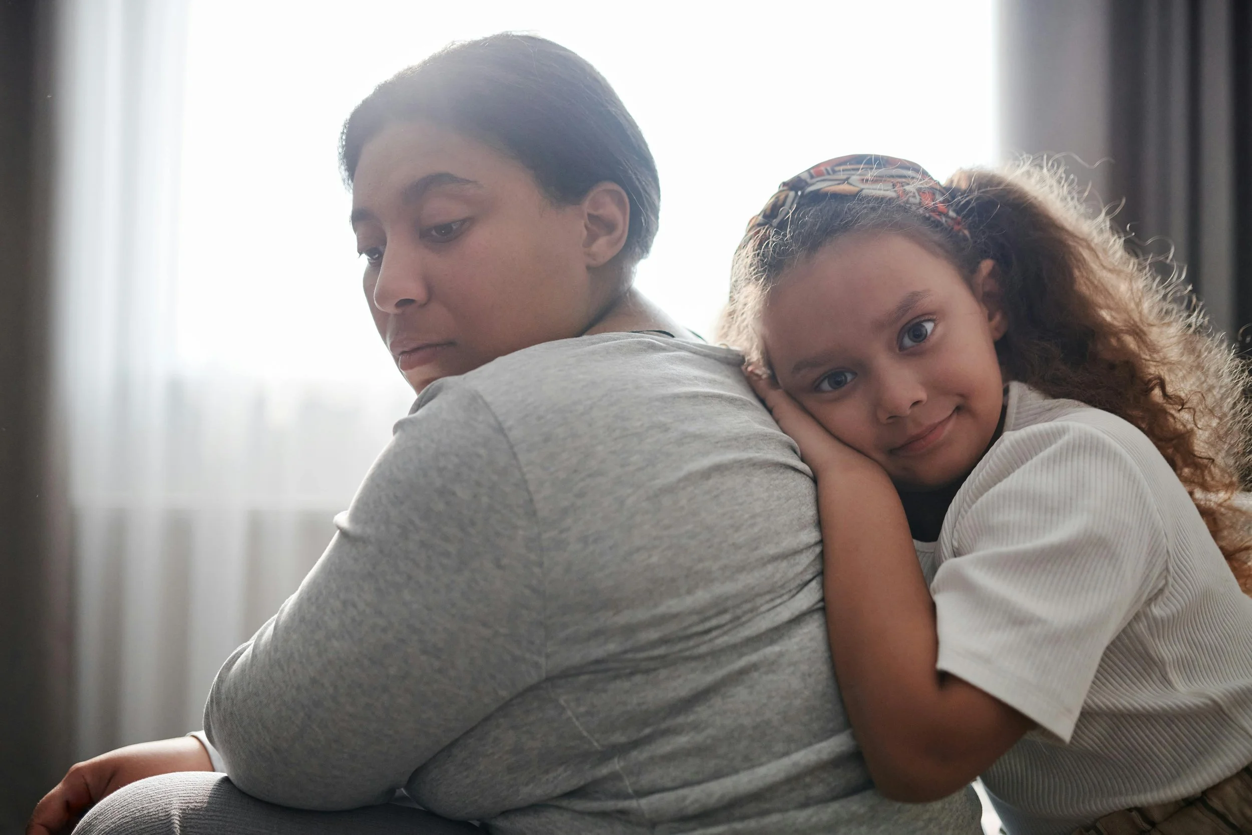 An exhausted mother sitting quietly while her young child leans affectionately against her back.