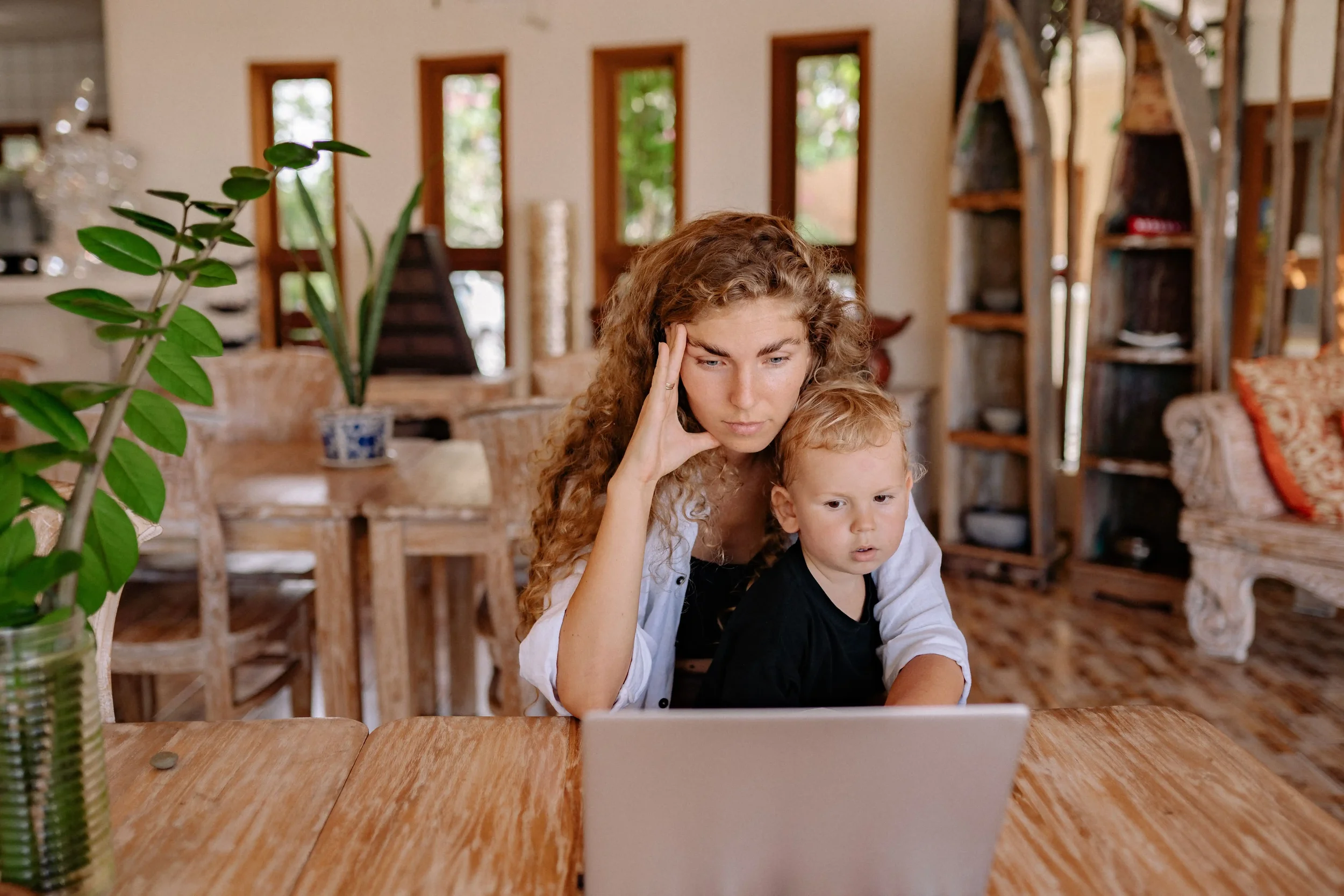 An overwhelmed mother looking at a laptop with her toddler, capturing the daily stress of juggling modern parenting pressure, work, and family schedules.