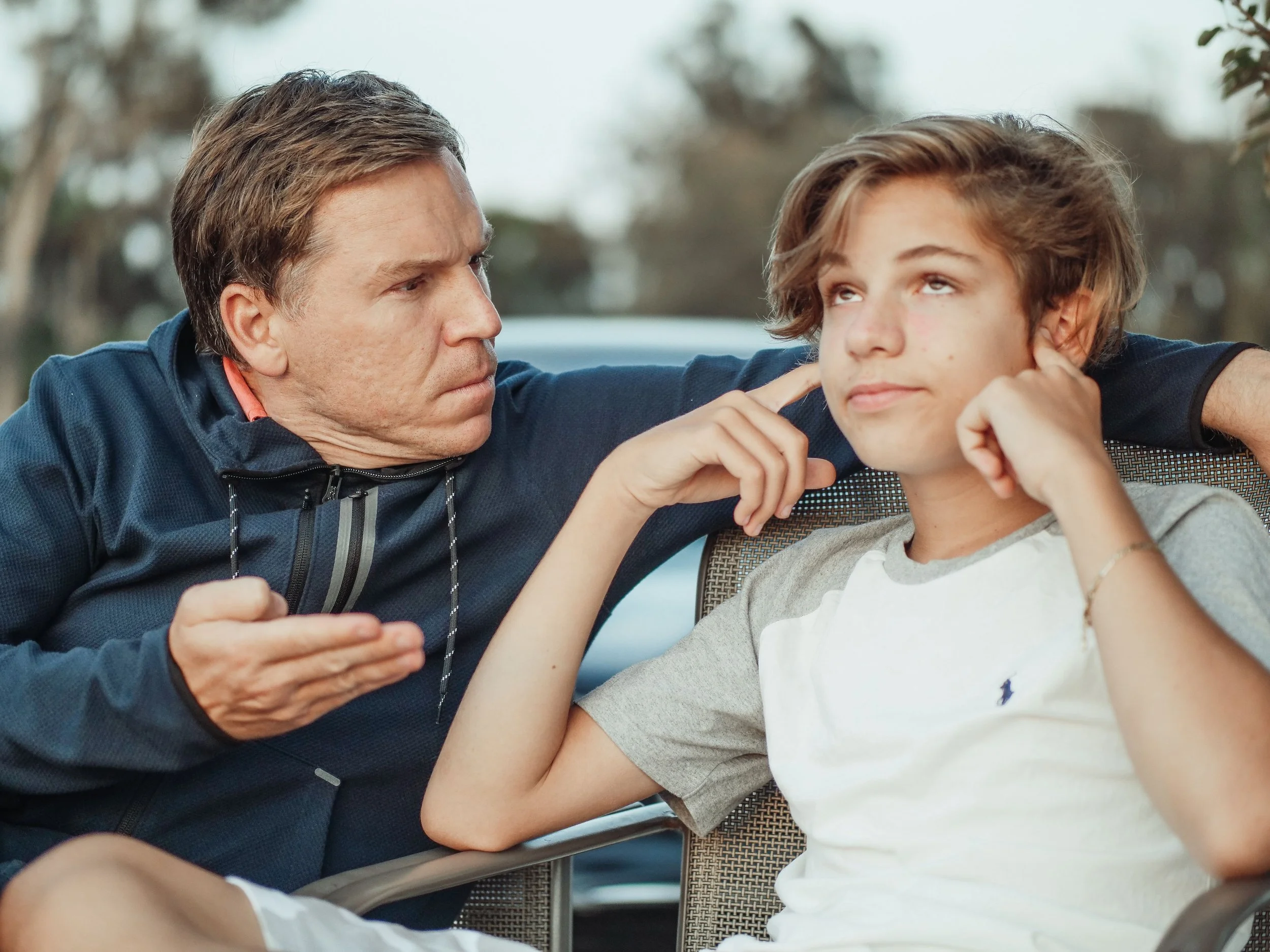 A frustrated father tries to talk to his teenage son, who is rolling his eyes and plugging his ears with his fingers.