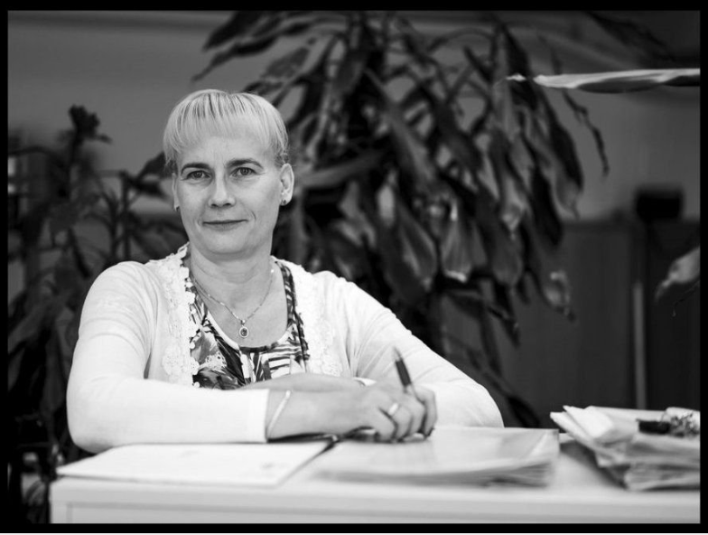 Nainen istuu pöydän ääressä kirjoituskoneen ja papereiden kanssa, taustalla huonekasveja.