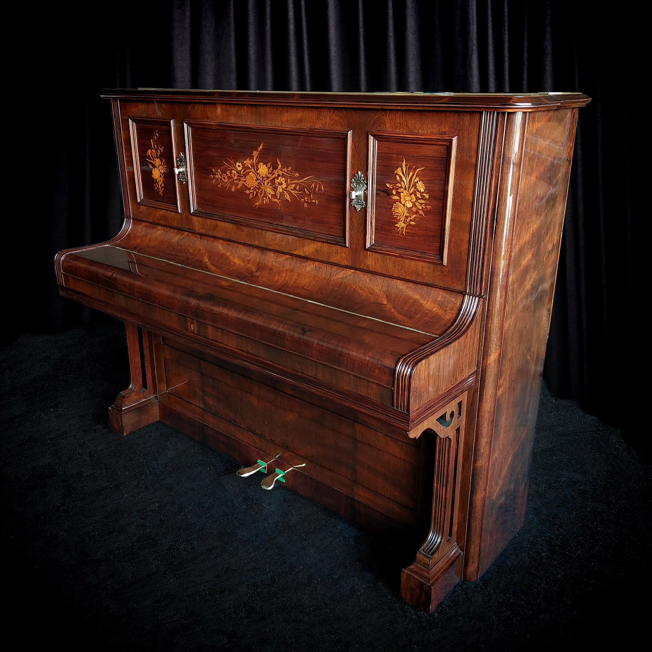 An antique upright piano made of polished dark wood with floral inlay on the front panel, set against a black curtain backdrop.
