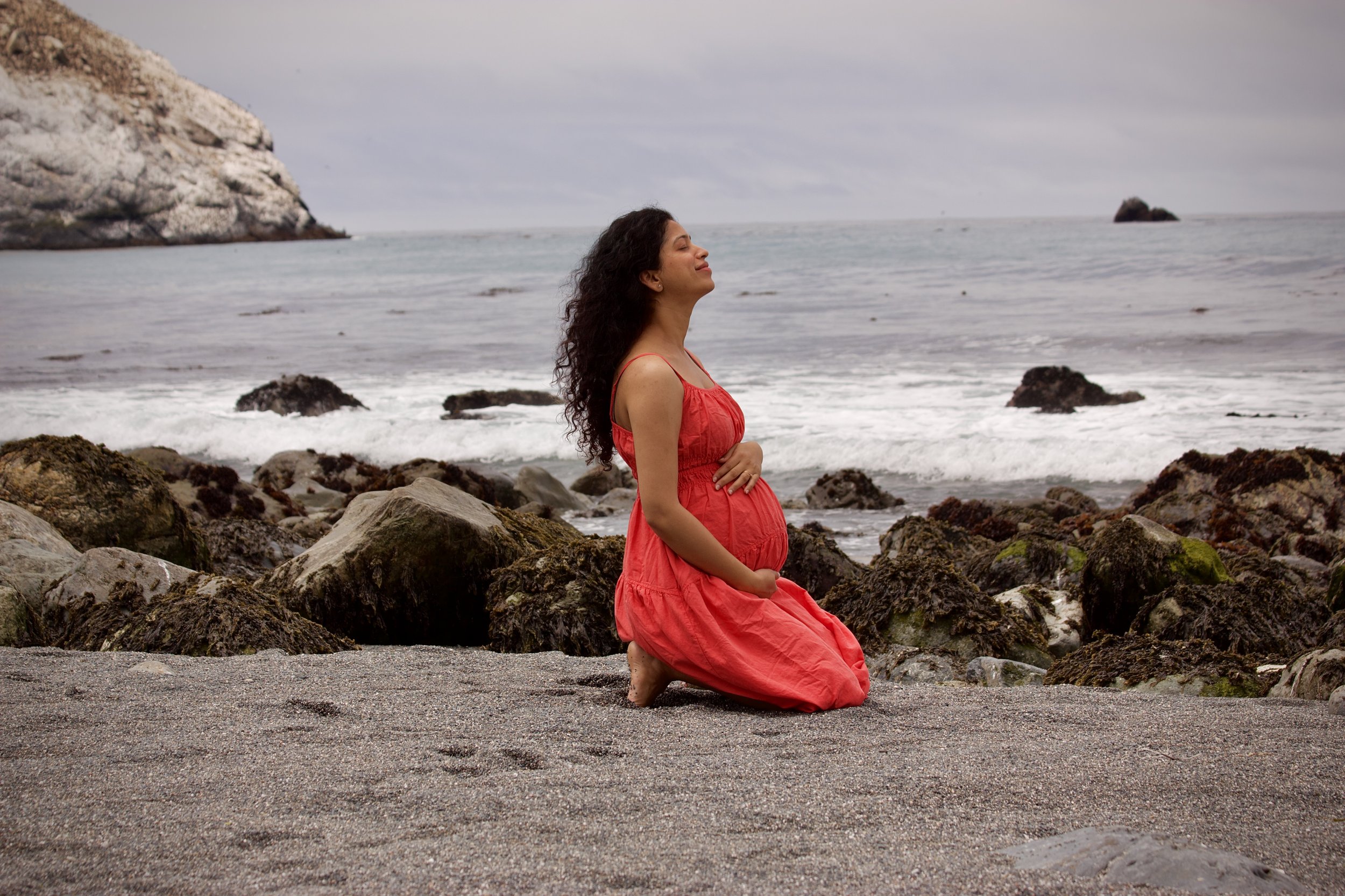 Pregnant woman in a red dress practising prenatal yoga on a beach, connecting with her body by the ocean