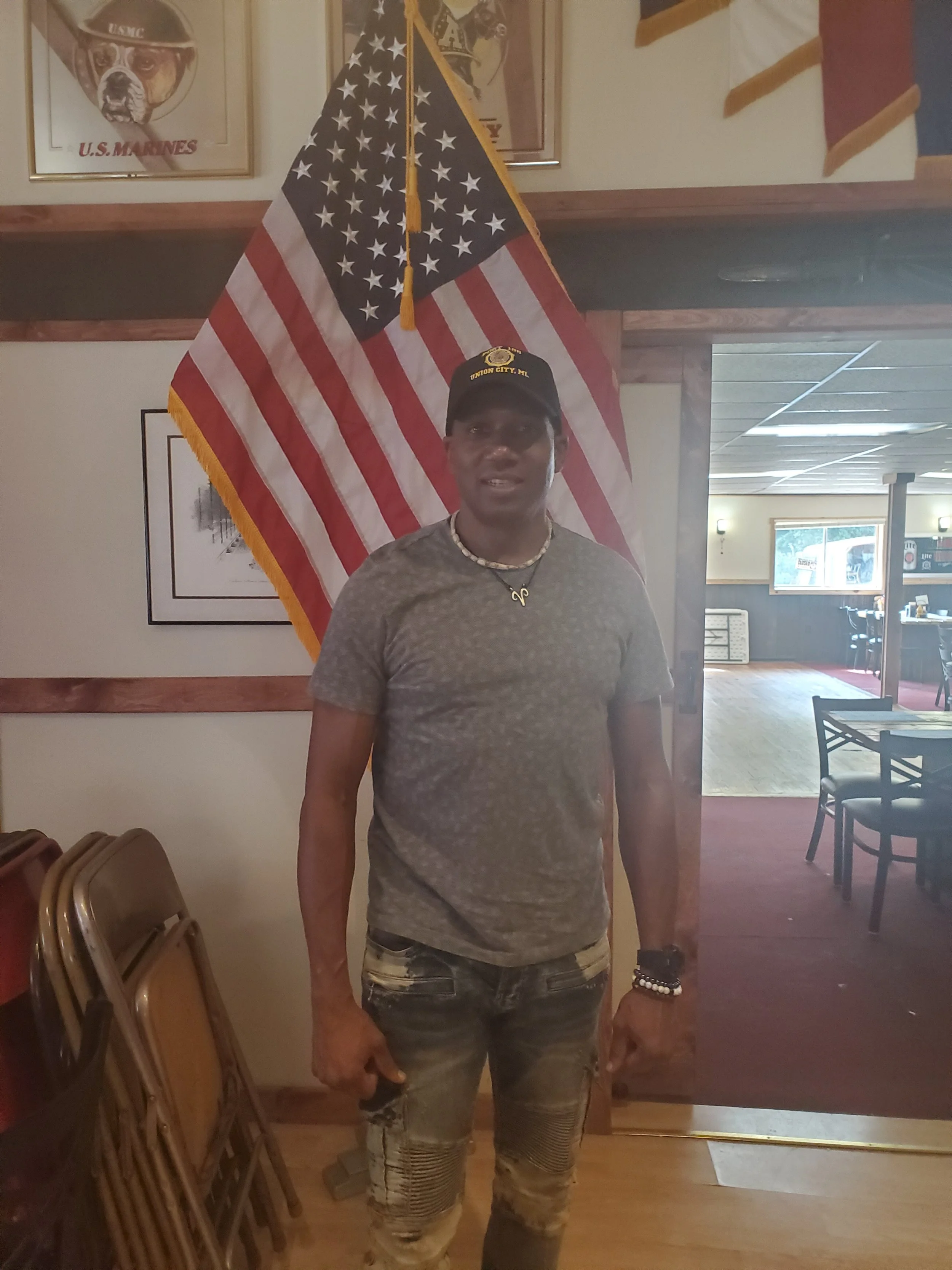 A man wearing a grey T-shirt, camouflage ripped jeans, a pearl necklace, and a black cap with yellow text stands in front of American and military flags inside a room with framed pictures on the wall and chairs stacked to the side.