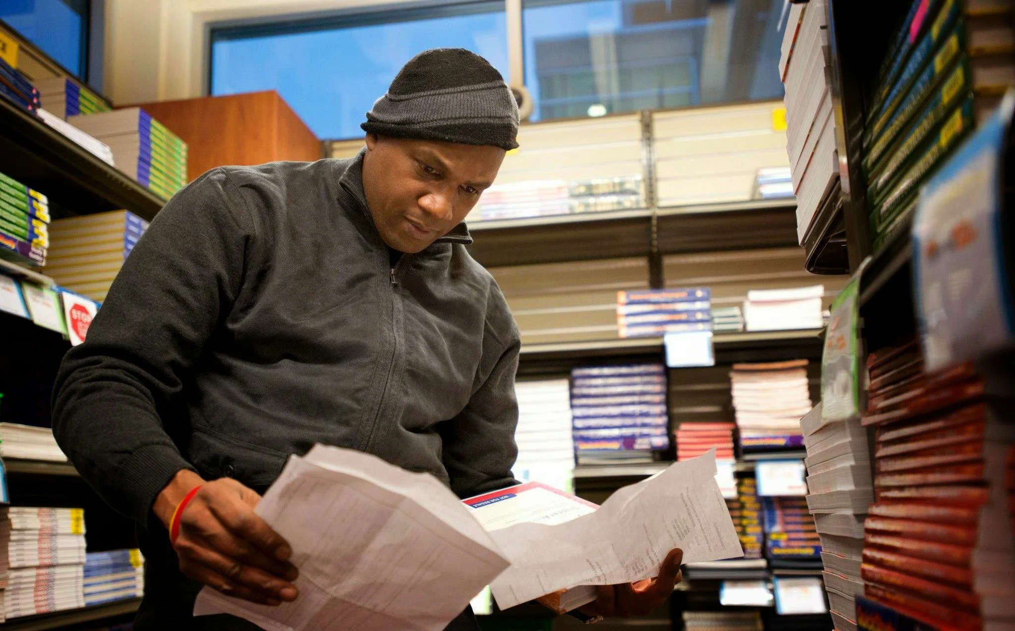 A man wearing a black beanie and jacket shopping for books in a bookstore, holding an open book in his hand.