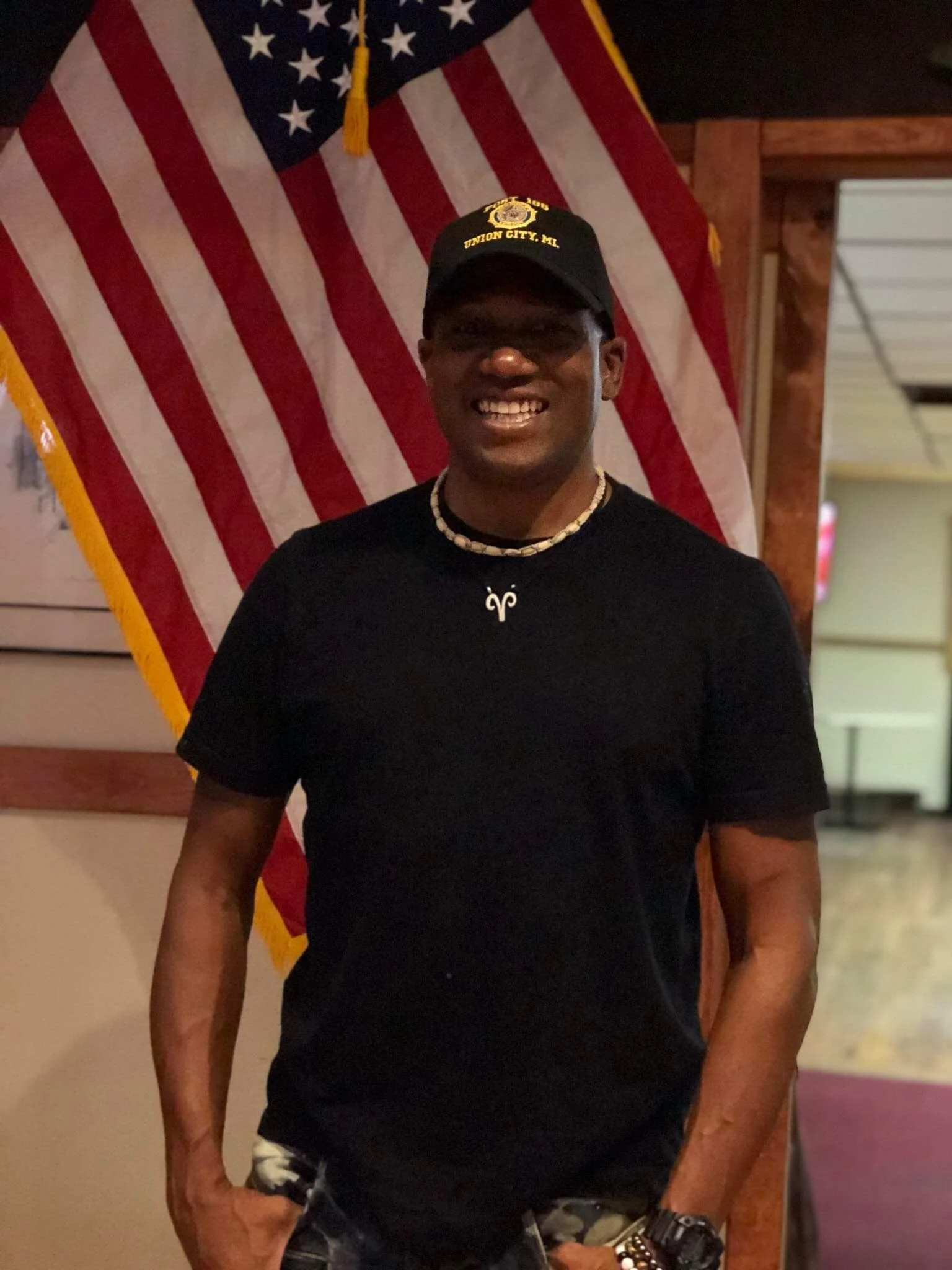 Man wearing a black t-shirt, camouflage shorts, a necklace, a watch, and bracelets, smiling in front of an American flag and a wooden wall.