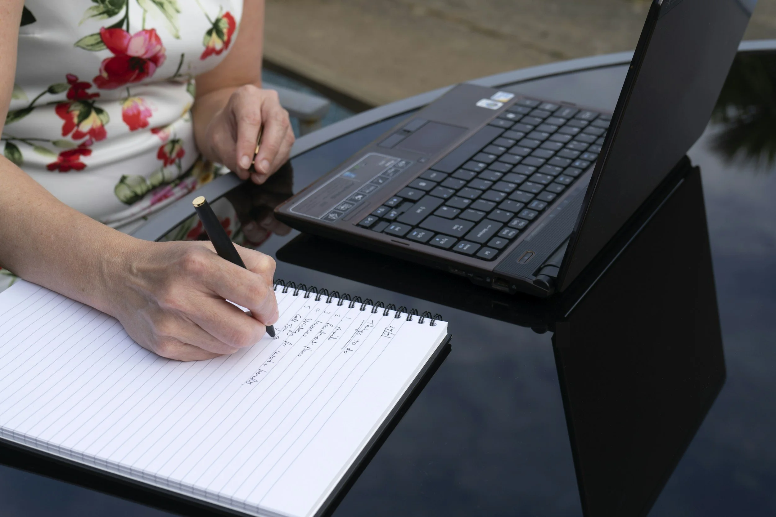 Person writing notes on a spiral notebook with a laptop on a black tabletop beside them.