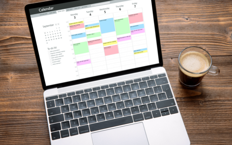 Laptop displaying a weekly digital calendar with scheduled events, next to a glass of coffee on a wooden table.