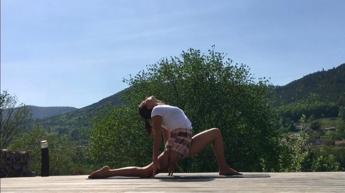 Eine Person macht Yoga im Freien auf einer Holzplattform vor Bergen und Bäumen.