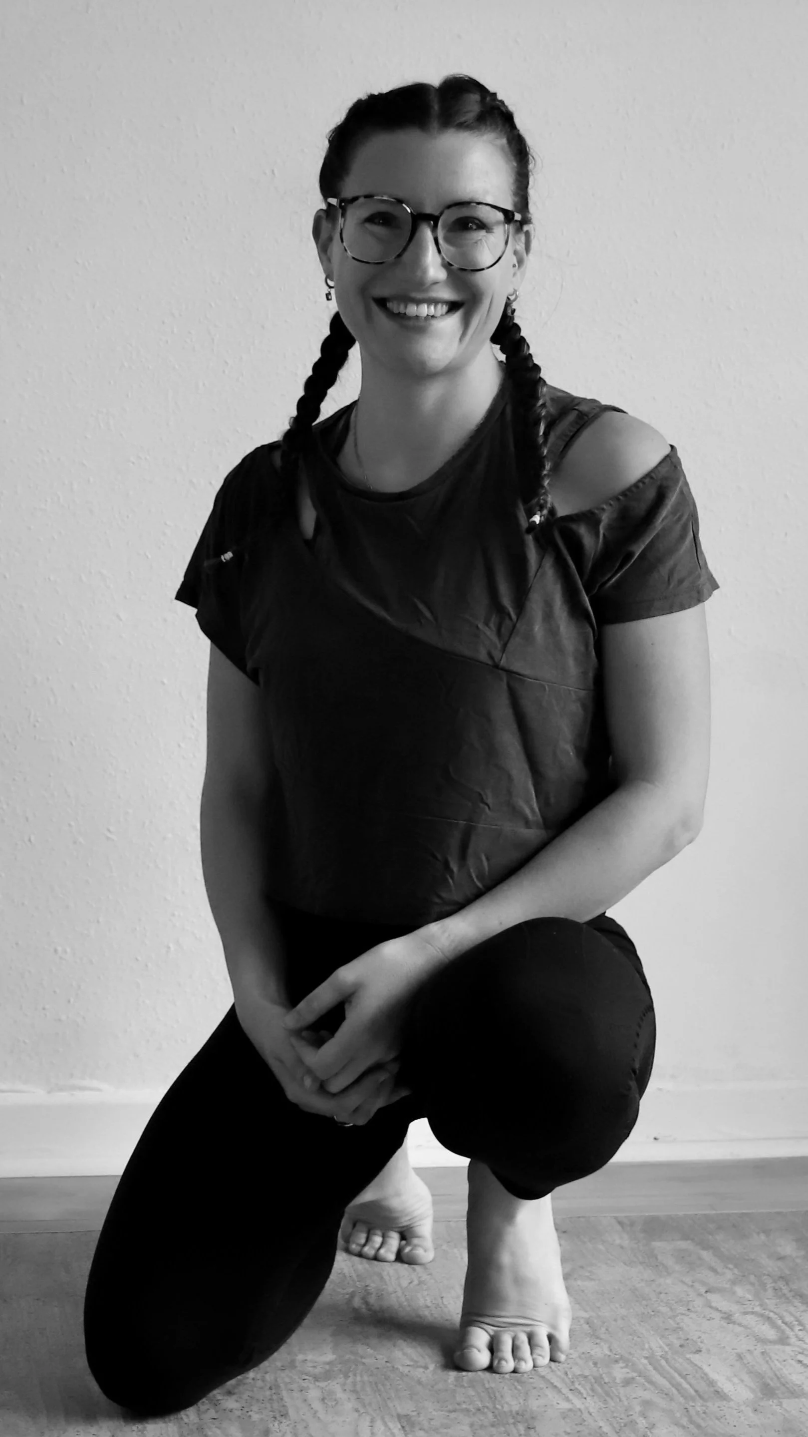 Schwarzweißfoto einer lachenden Frau mit Brille, die in einer Yoga-Pose auf einem Fuß und Knie an einem Holzboden auf einem weißen Wand steht.