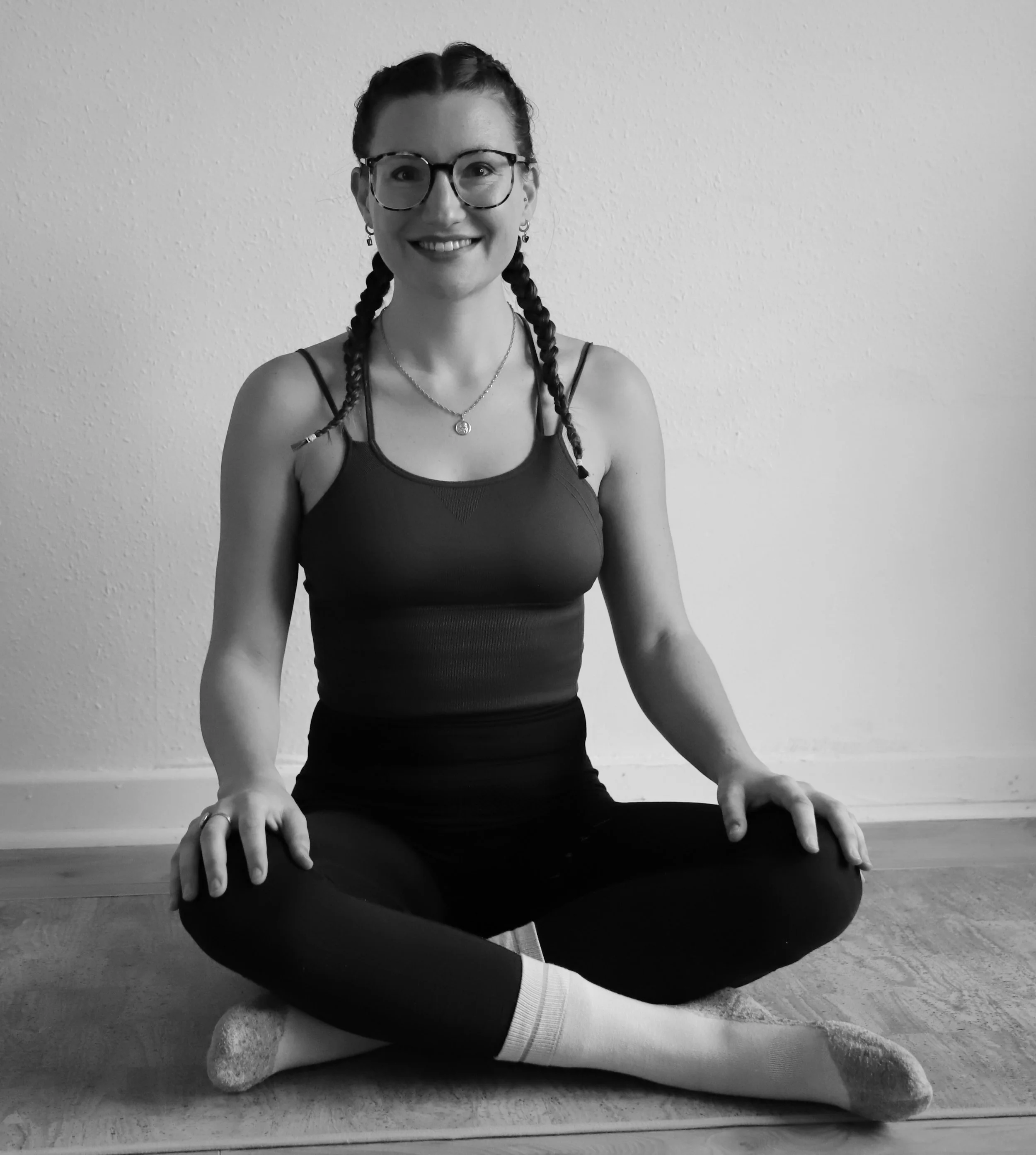 Eine lächelnde junge Frau mit Brille und Zöpfen macht Yoga im Sitzen auf einem Boden, mit eingerolltem Handtuch unter den Knien, vor einer weißen Wand.