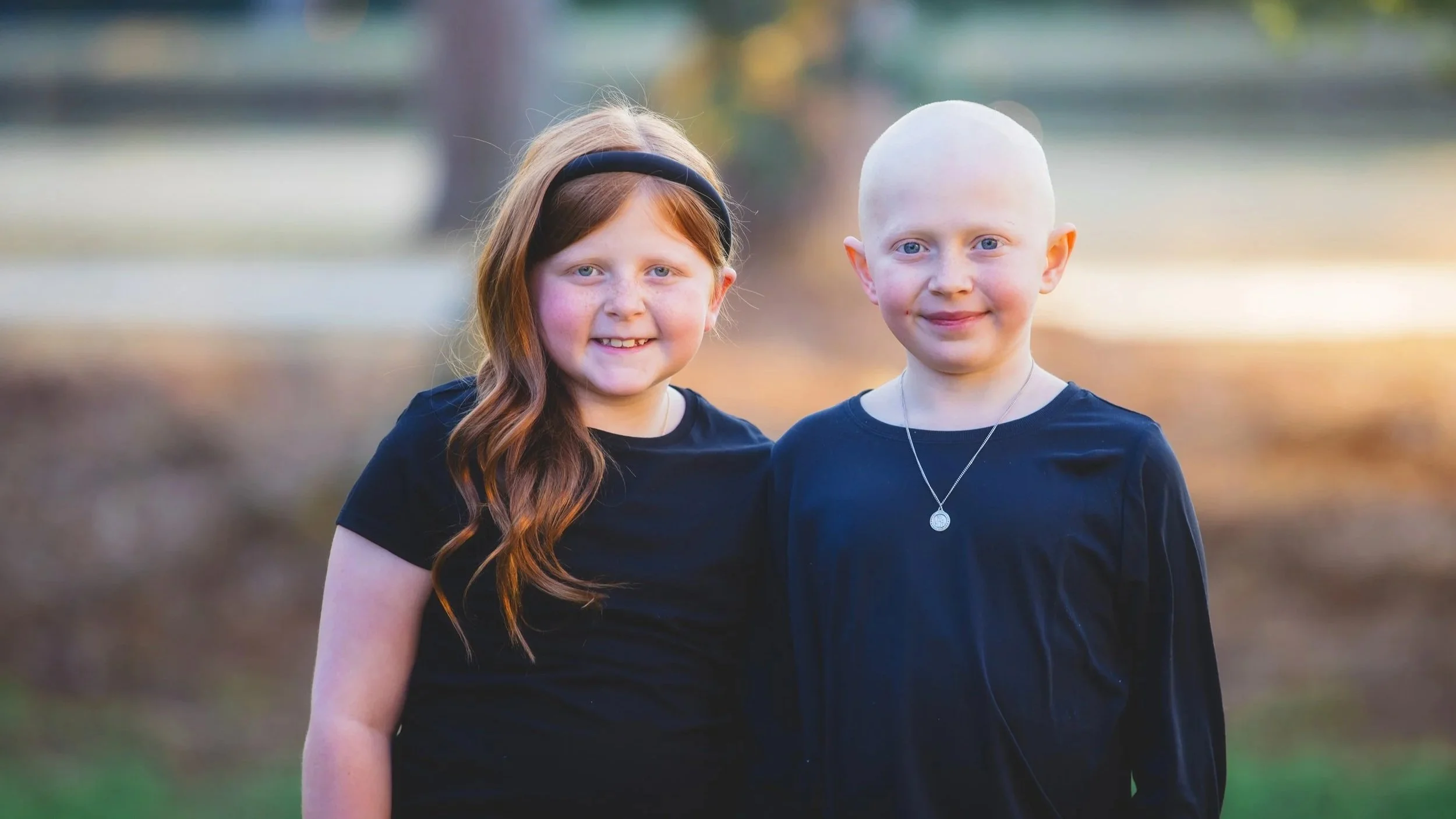 Two young girls standing outdoors, smiling, dressed in black, with trees and a blurred background behind them.