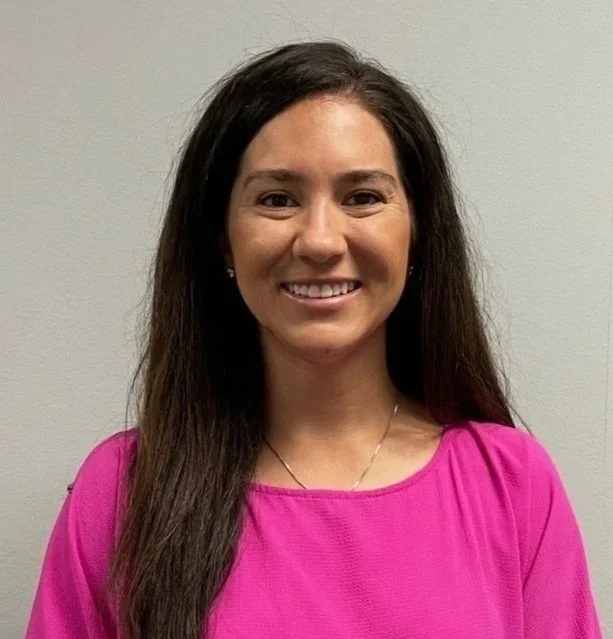 A young woman with long dark hair and light brown skin smiling, wearing a bright pink top and small earrings, standing against a plain light-colored background.