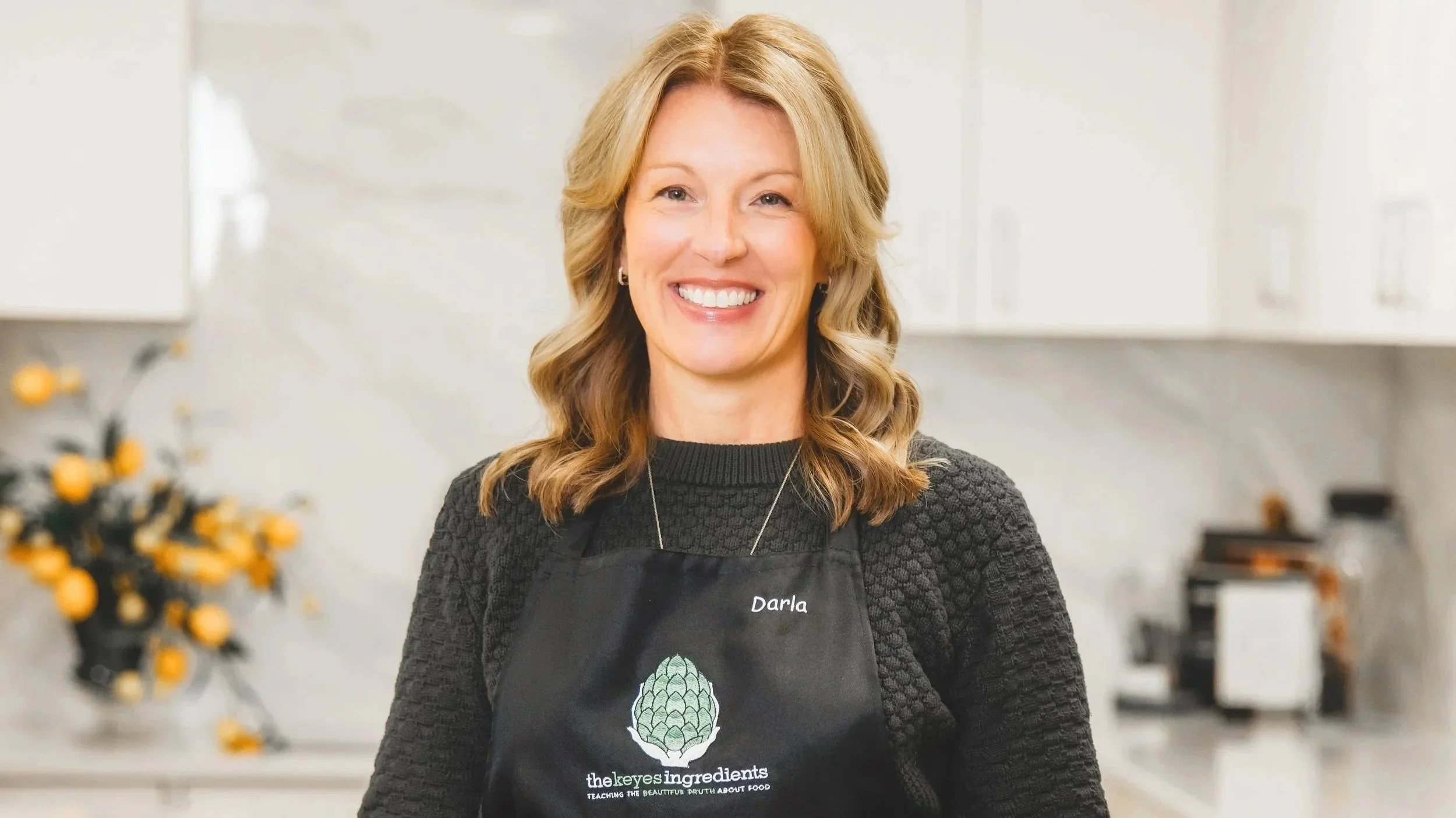 A woman with shoulder-length blond hair, smiling, wearing a black apron with the word Darla and a logo of an artichoke, standing in a kitchen.