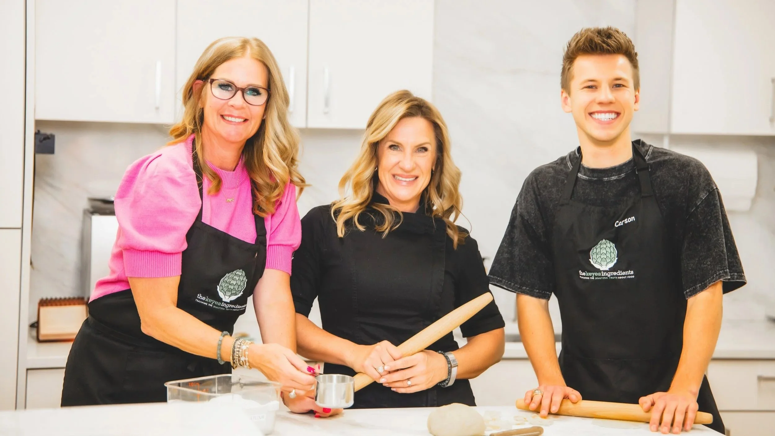 Three women and one young man smiling and posing in a kitchen, wearing black aprons, with rolling pins and baking supplies on the counter.