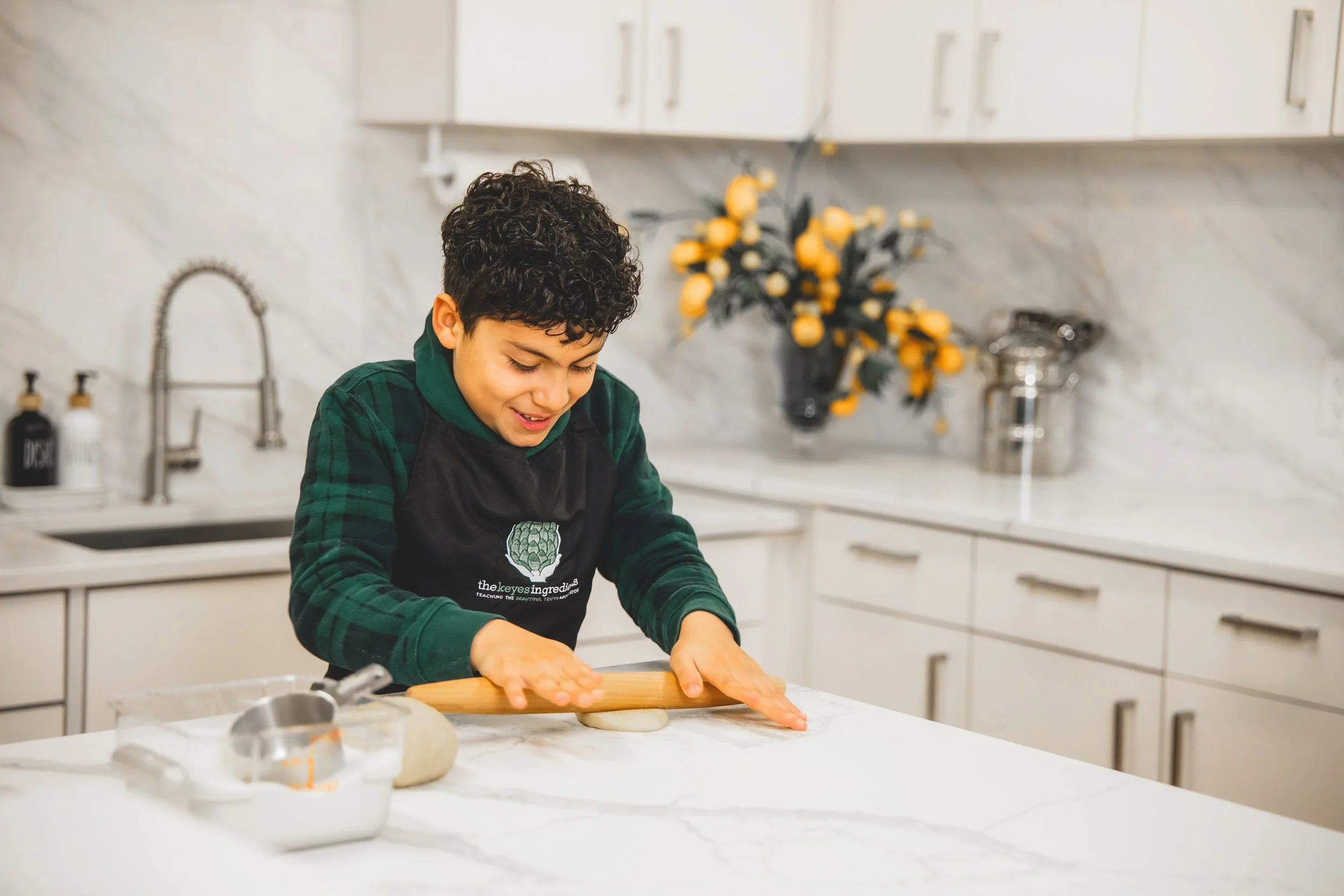 A young boy in a kitchen, rolling dough on a white marble countertop. He's smiling and wearing a black apron and a green and black plaid shirt. There is a vase of yellow flowers and kitchen cabinets in the background.