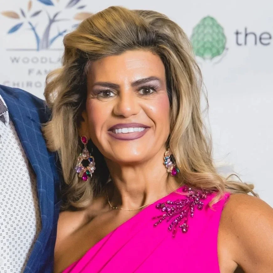 A woman with blonde hair styled in loose curls, wearing a bright pink dress with embellished details on the shoulder. She is smiling and wearing colorful earrings and a delicate necklace.