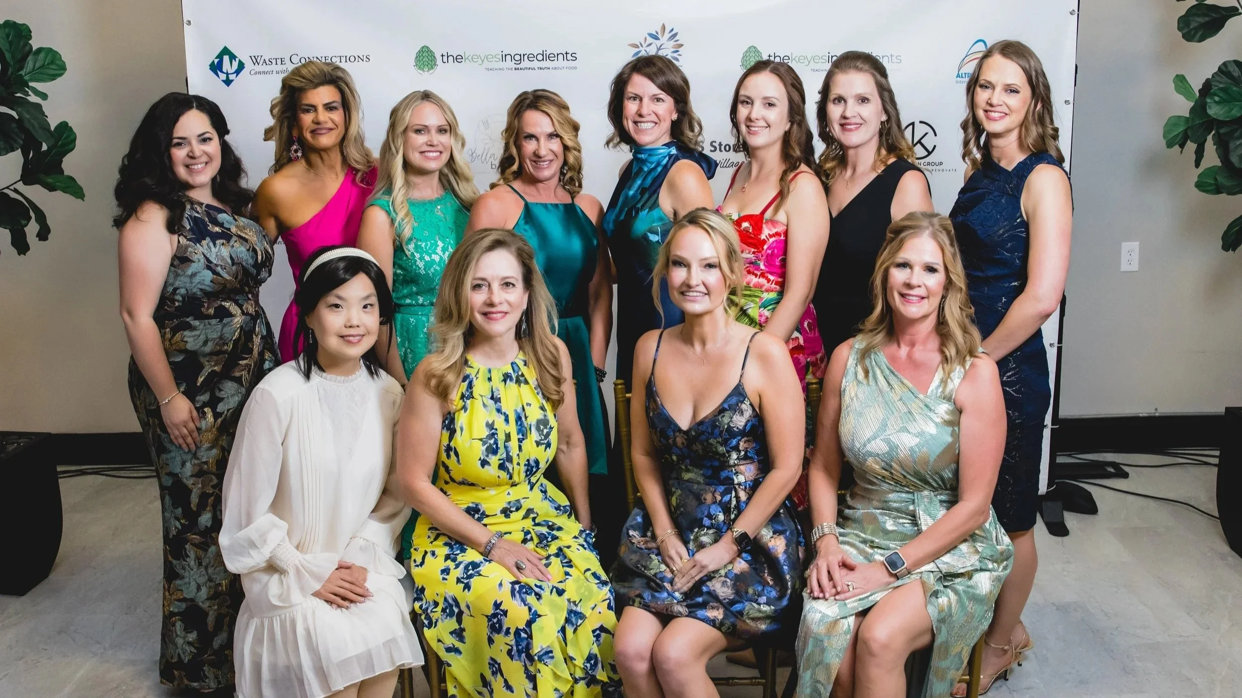 A group of fourteen women dressed in colorful dresses posing for a photo indoors in front of a backdrop with logos, including Waste Connections and the keyesingredients.