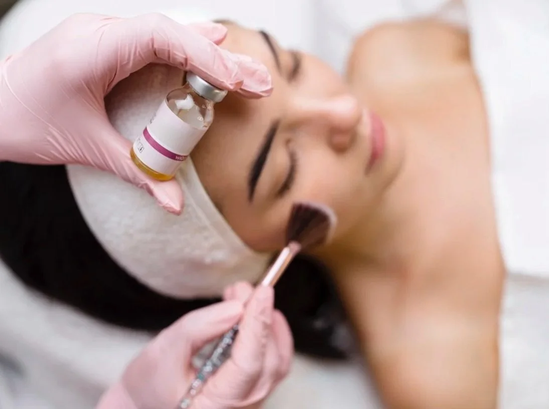 Eine Frau wird in einem Schönheitsstudio mit einer Gesichtsschutzmaske und einer Vlieshaube behandelt, während eine Kosmetikerin eine Botox-Injektion verabreicht und Make-up aufträgt.