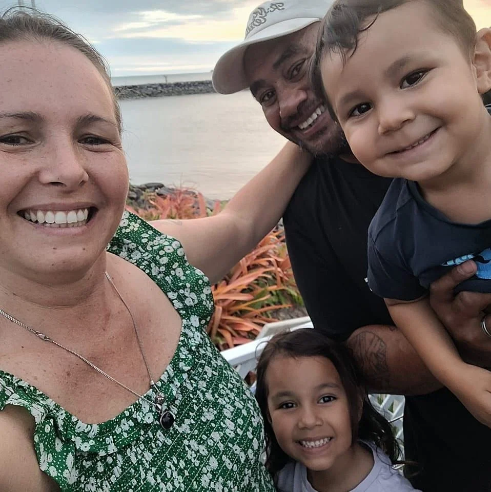 A happy family of four taking a selfie outdoors near a body of water with a cloudy sky.