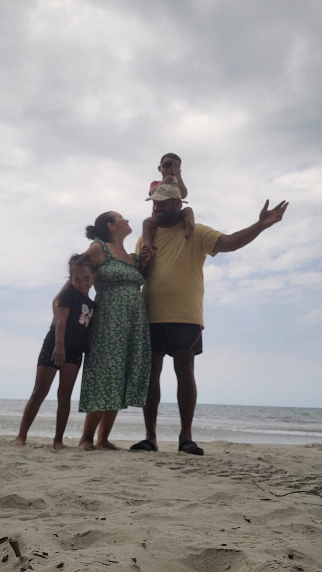 A family of four, including a man, woman, and two children, on the beach. The man is holding one child on his shoulders, and the woman is standing beside him, while the other child is leaning against her. They are all enjoying a moment together with the ocean and cloudy sky in the background.