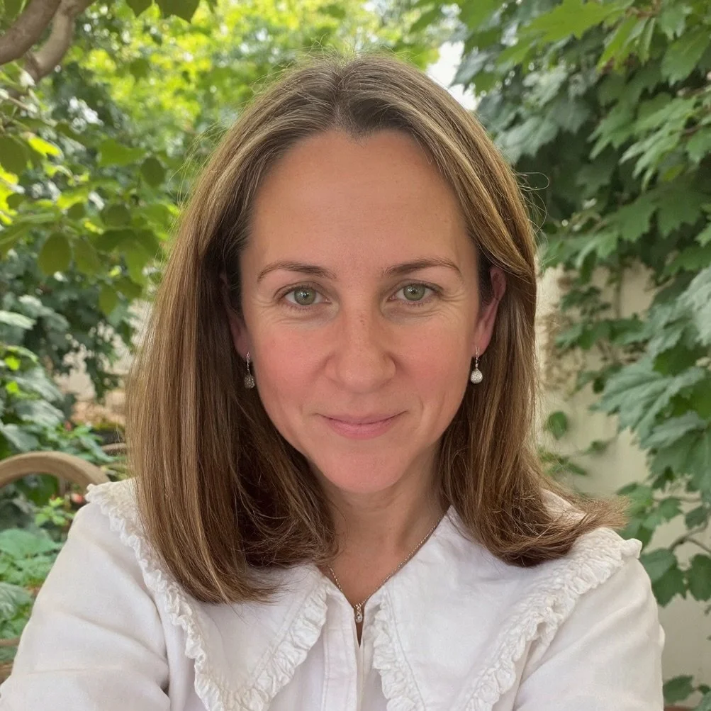 A woman with brown hair and green eyes smiling outdoors with green leafy plants in the background.