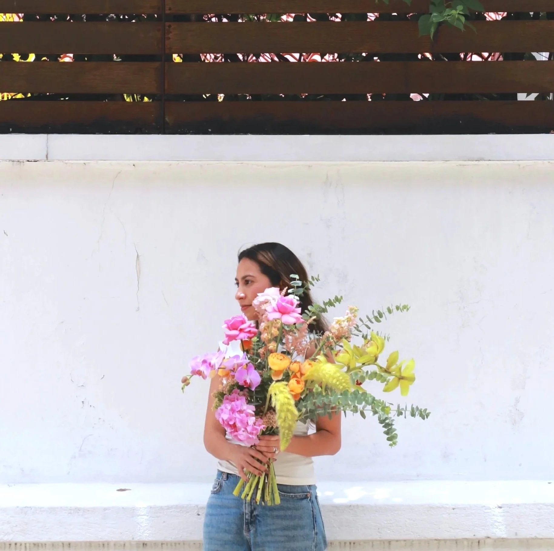 Woman holding a large bouquet of colorful flowers in front of a white wall and brown slatted fence.