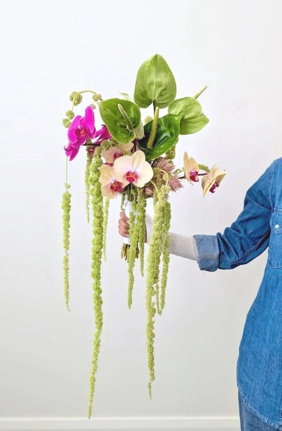 Hand holding a tall floral arrangement with pink, white, and purple orchids and long, hanging green amaranthus.
