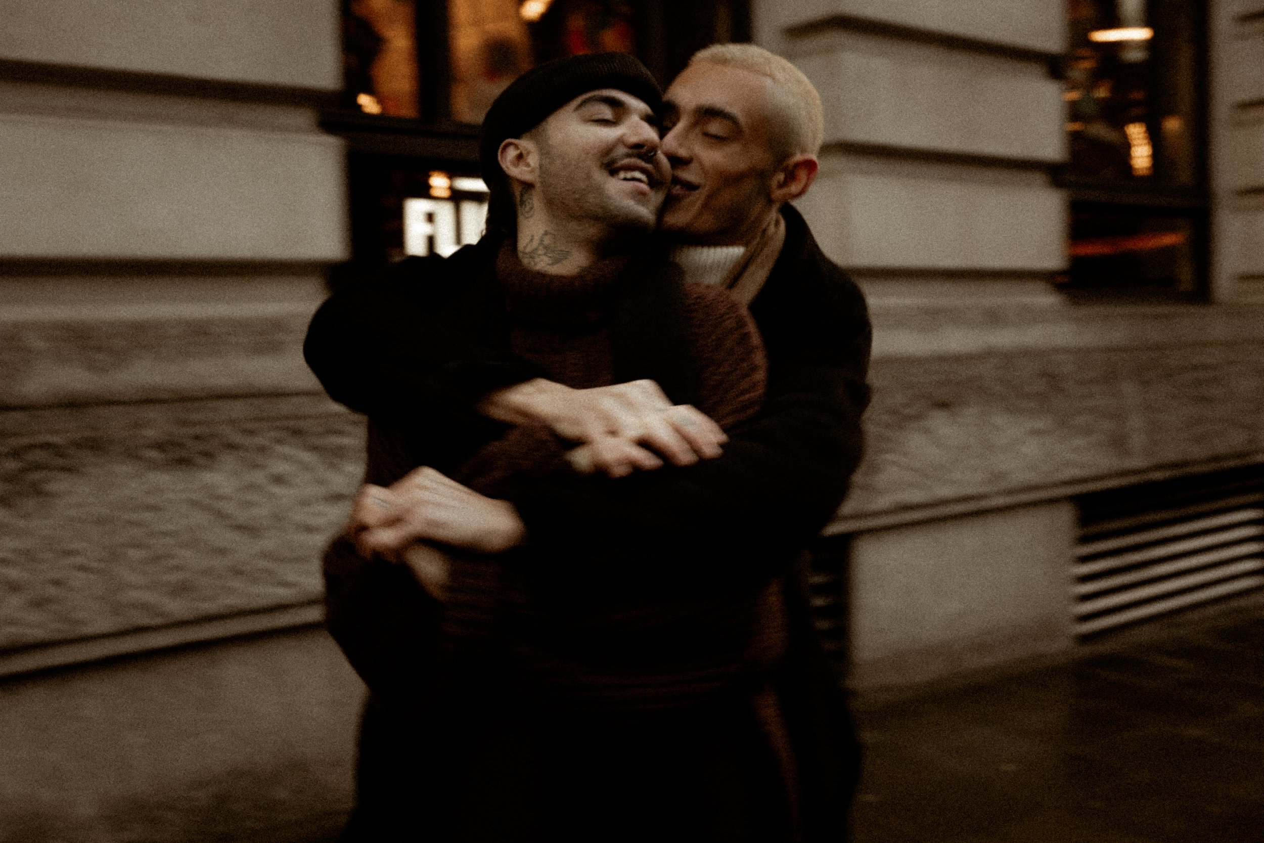 Two men embracing and sharing a kiss on a city sidewalk at night.
