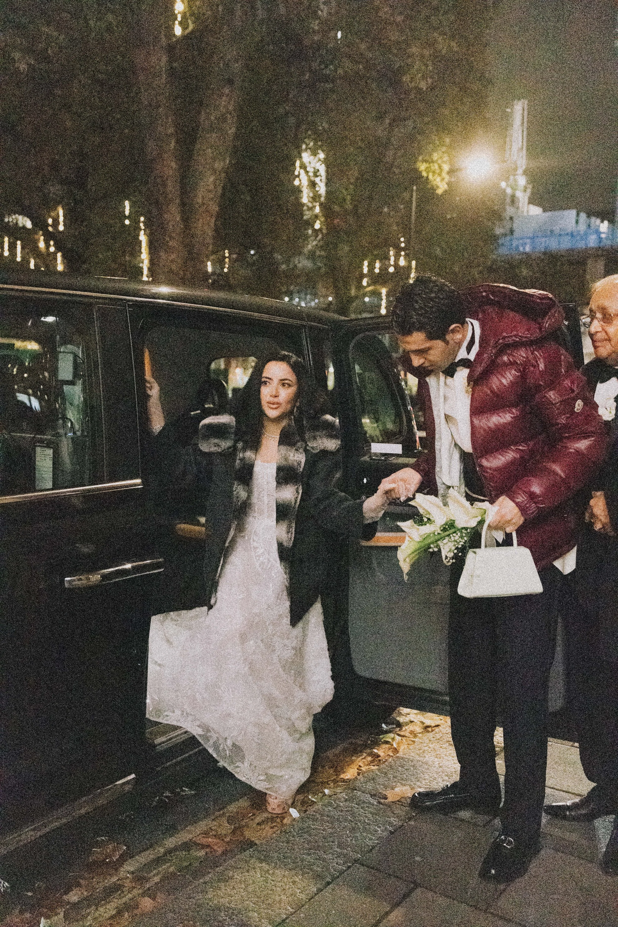 A woman in a white wedding dress is stepping out of a black vehicle, being handed a bouquet of flowers by a man in a maroon puffer jacket, with another man beside them, on a city street at night.