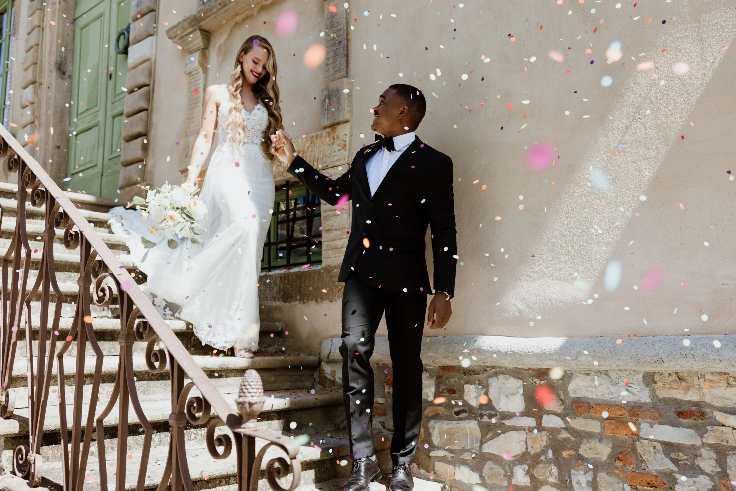 A newlywed couple celebrating on steps with falling confetti. The bride is holding a bouquet, wearing a white wedding dress, and smiling. The groom is dressed in a black tuxedo with a bow tie, holding the bride's hand and smiling at her.