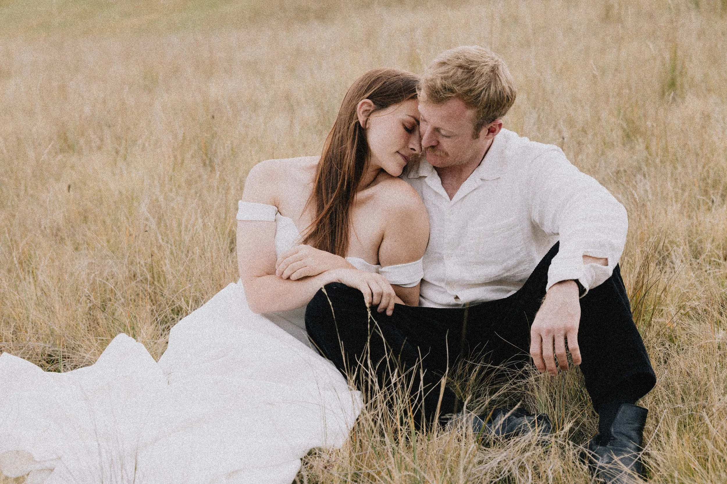 A couple sitting close together in a grassy field, with their foreheads touching and eyes closed. The woman is wearing an off-shoulder white dress, and the man is dressed in a white shirt and black pants.