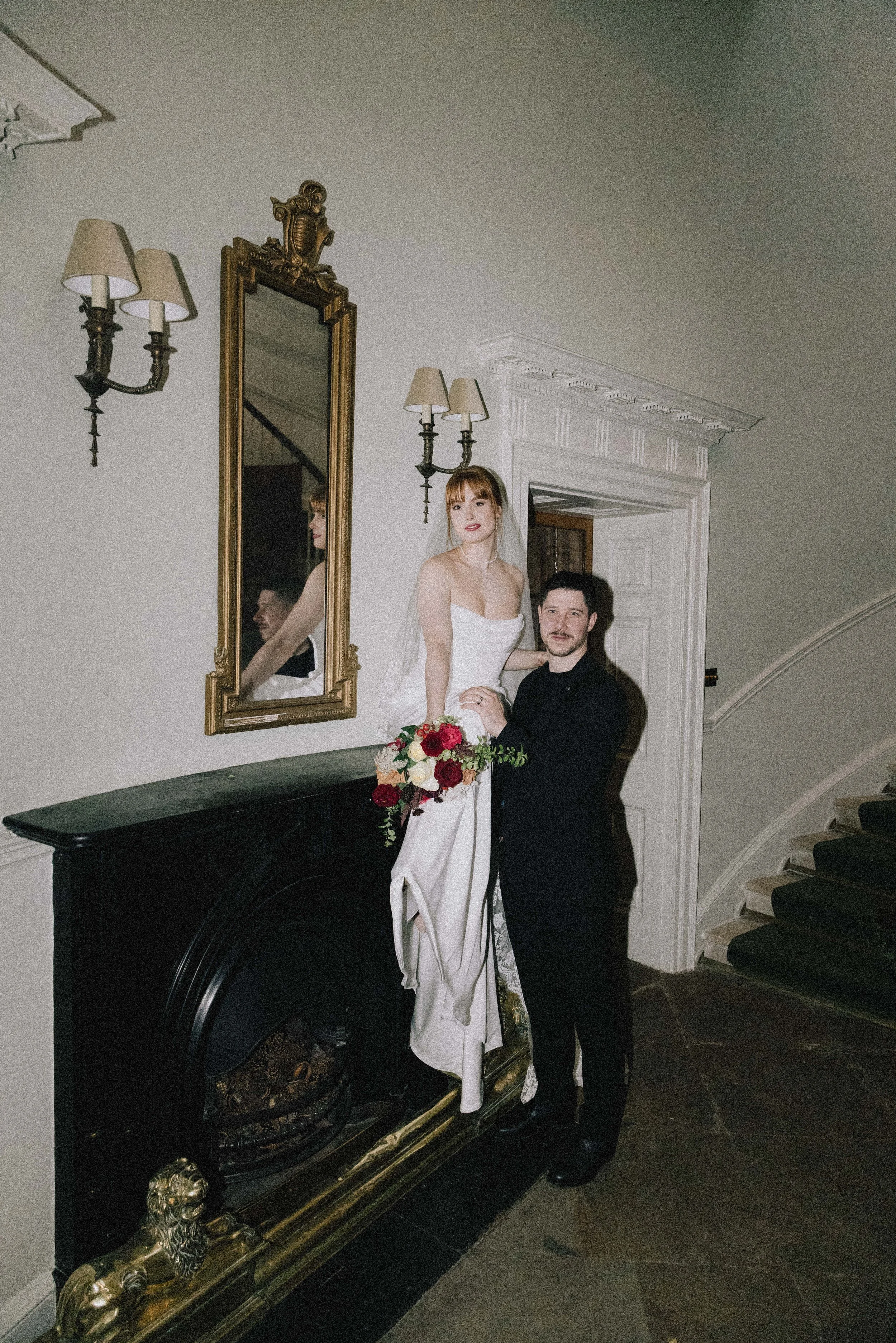 A bride in a white wedding dress holding a bouquet of red and white flowers stands next to a groom in a black suit, both posing inside a decorated room with a mirror, wall sconces, and a staircase.