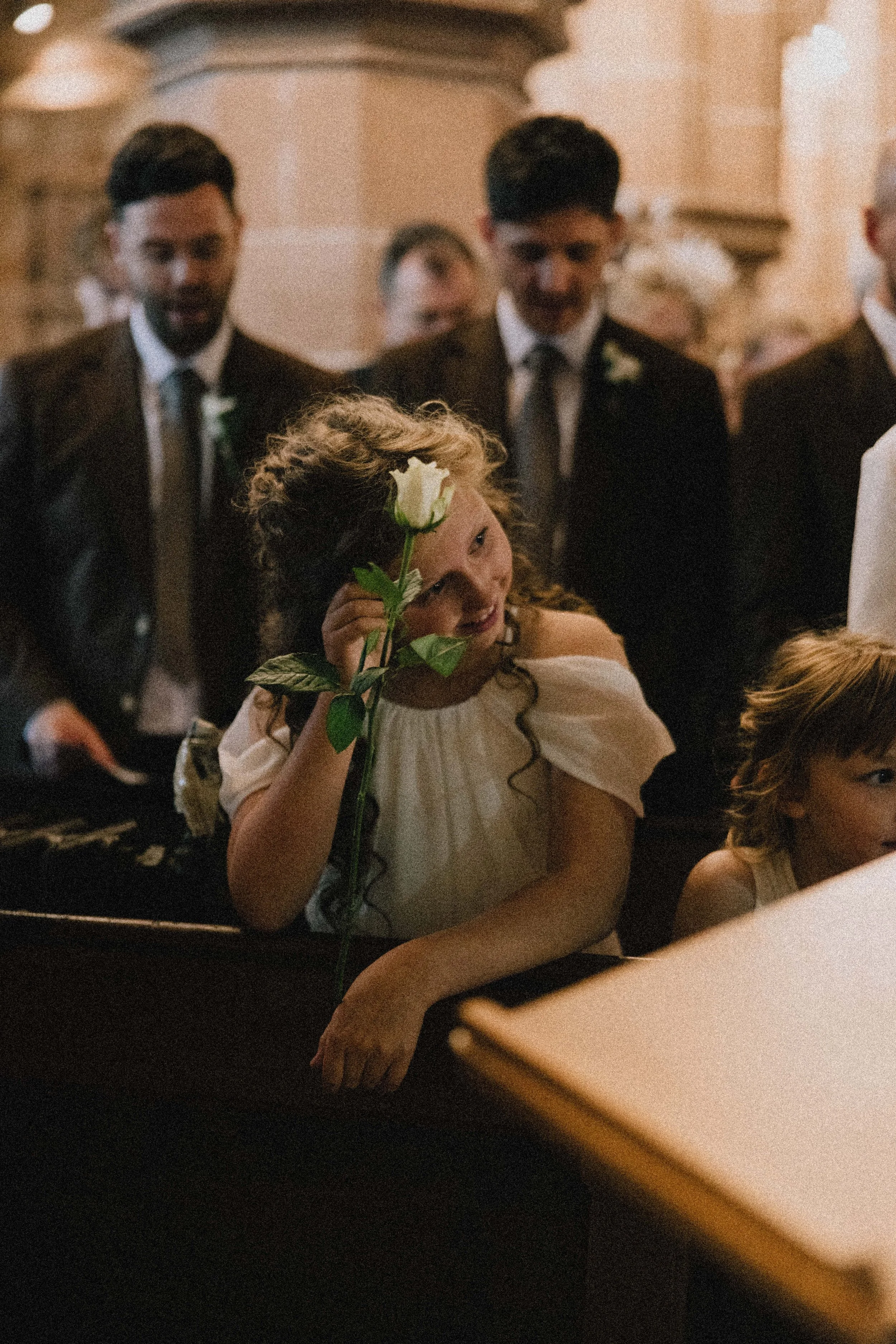 A girl with curly hair holding a white rose near her face in a church, surrounded by men in suits
