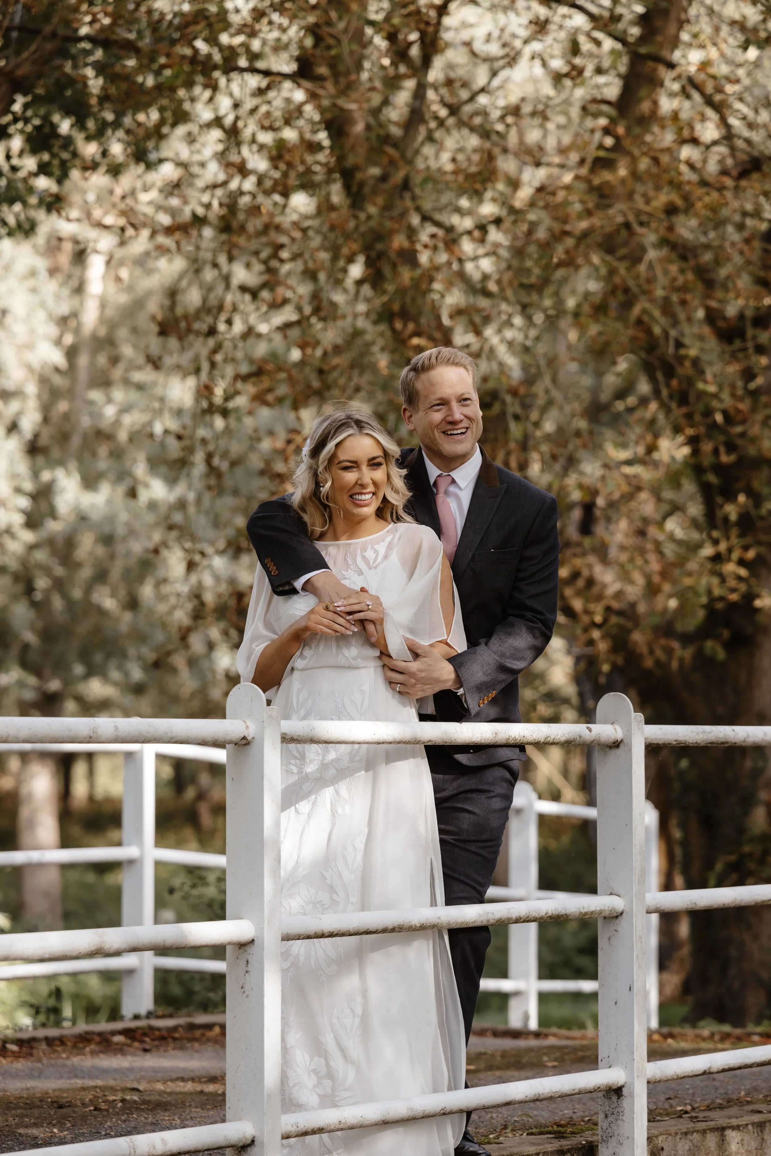 A smiling bride and groom standing together outdoors behind a white fence, embracing each other, with trees in the background. wedding at rowton castle 