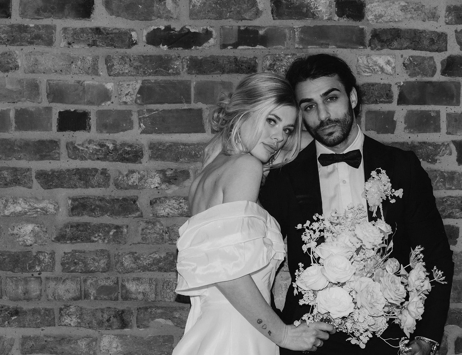 A black-and-white photo of a bride and groom posing together in front of a brick wall, with the bride holding a bouquet of flowers.