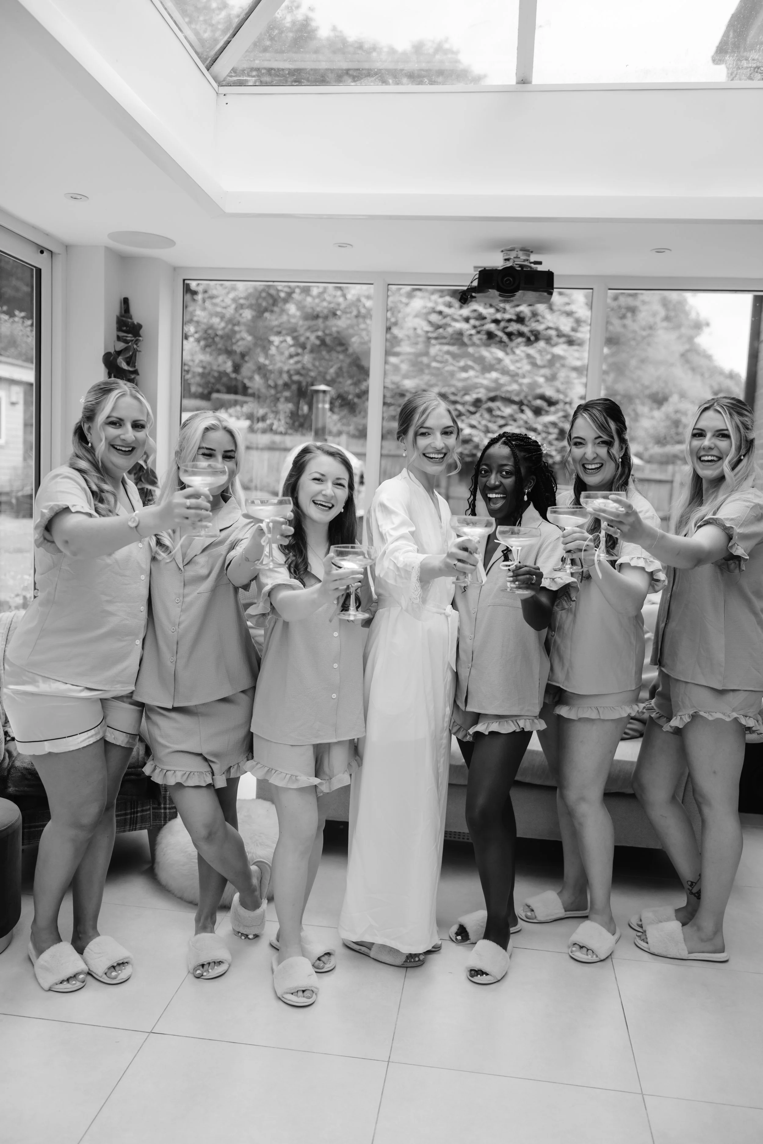 A group of eight women, dressed in pajamas, celebrating and holding glasses of champagne in a bright living room with large windows. One woman in the center wears a white dress, possibly indicating a bridal celebration. documentary wedding photograph