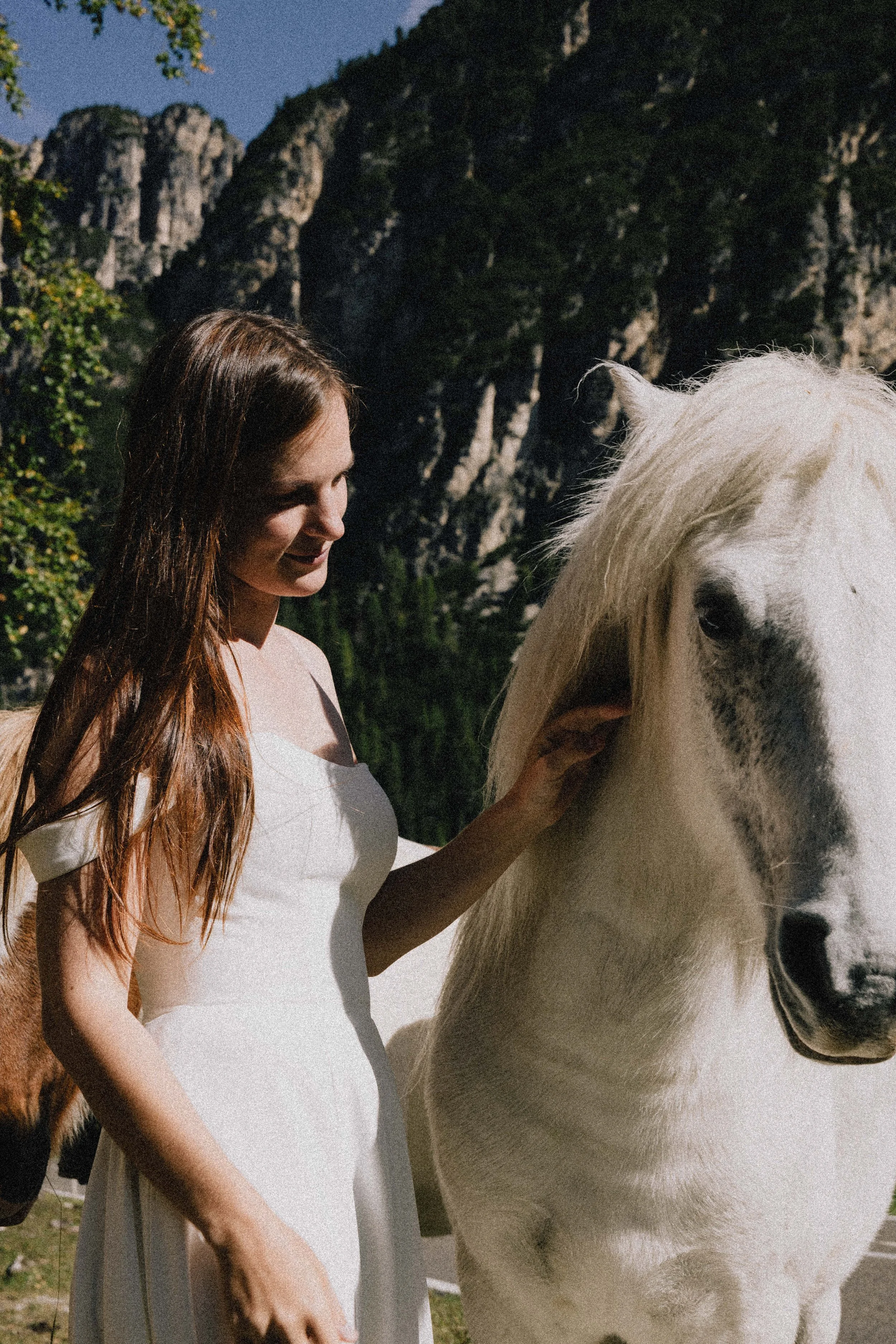 A woman with long brown hair in a white dress petting a white horse with black markings near a mountainous area.