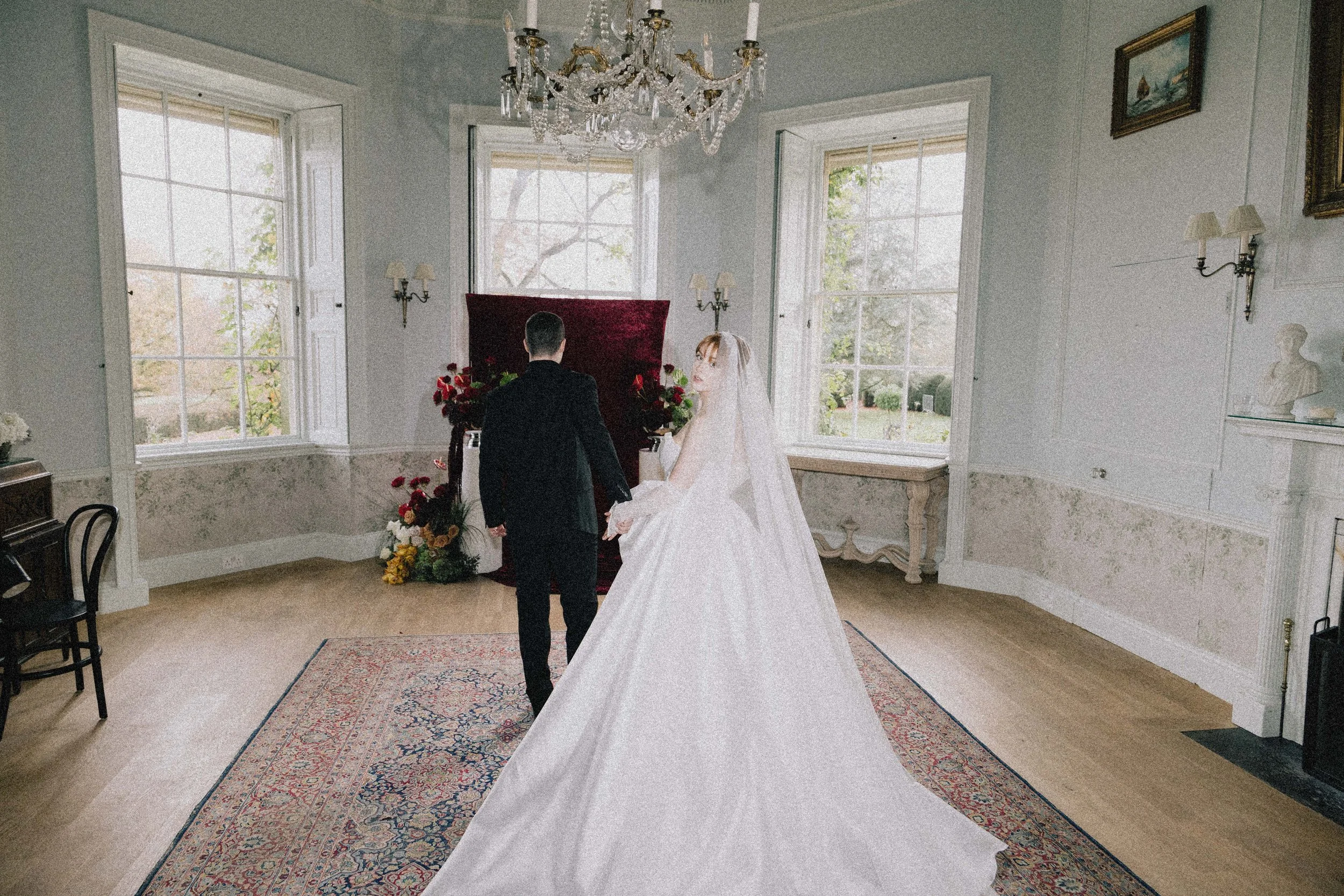 A bride and groom holding hands in a room with large windows, a chandelier, and traditional decor, during their wedding ceremony.