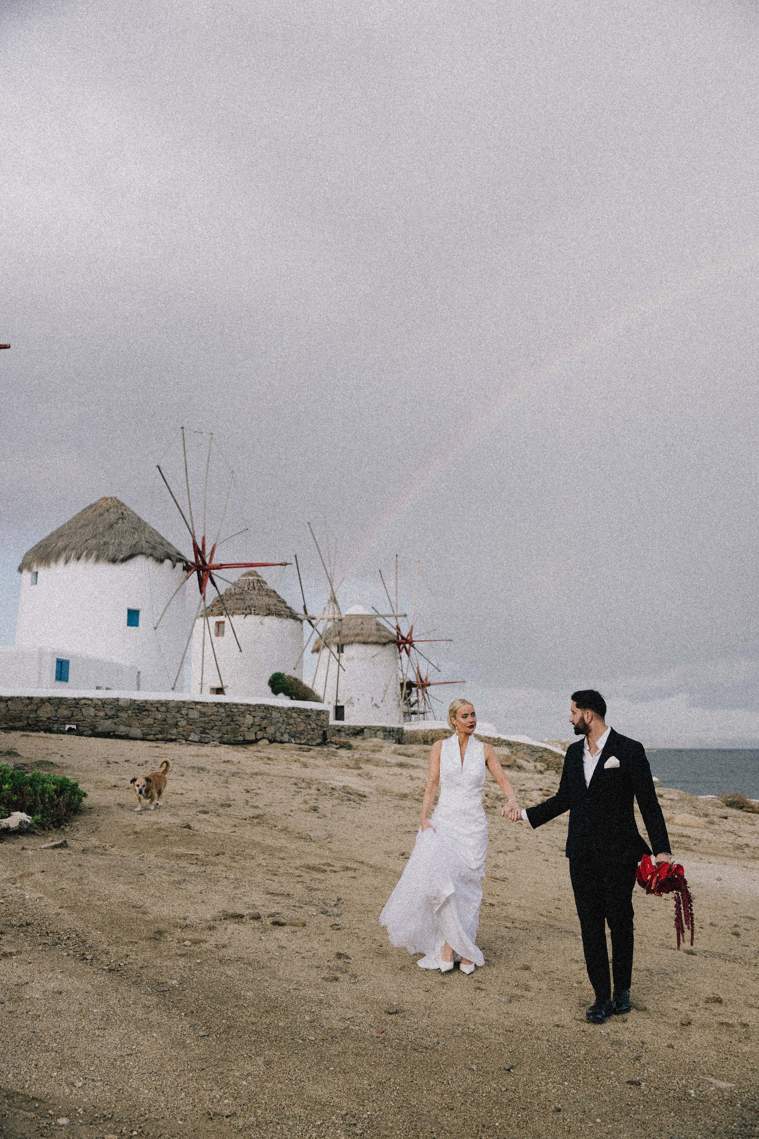 Couple in wedding attire holding hands on a beach with traditional windmills in the background and a cloudy sky.