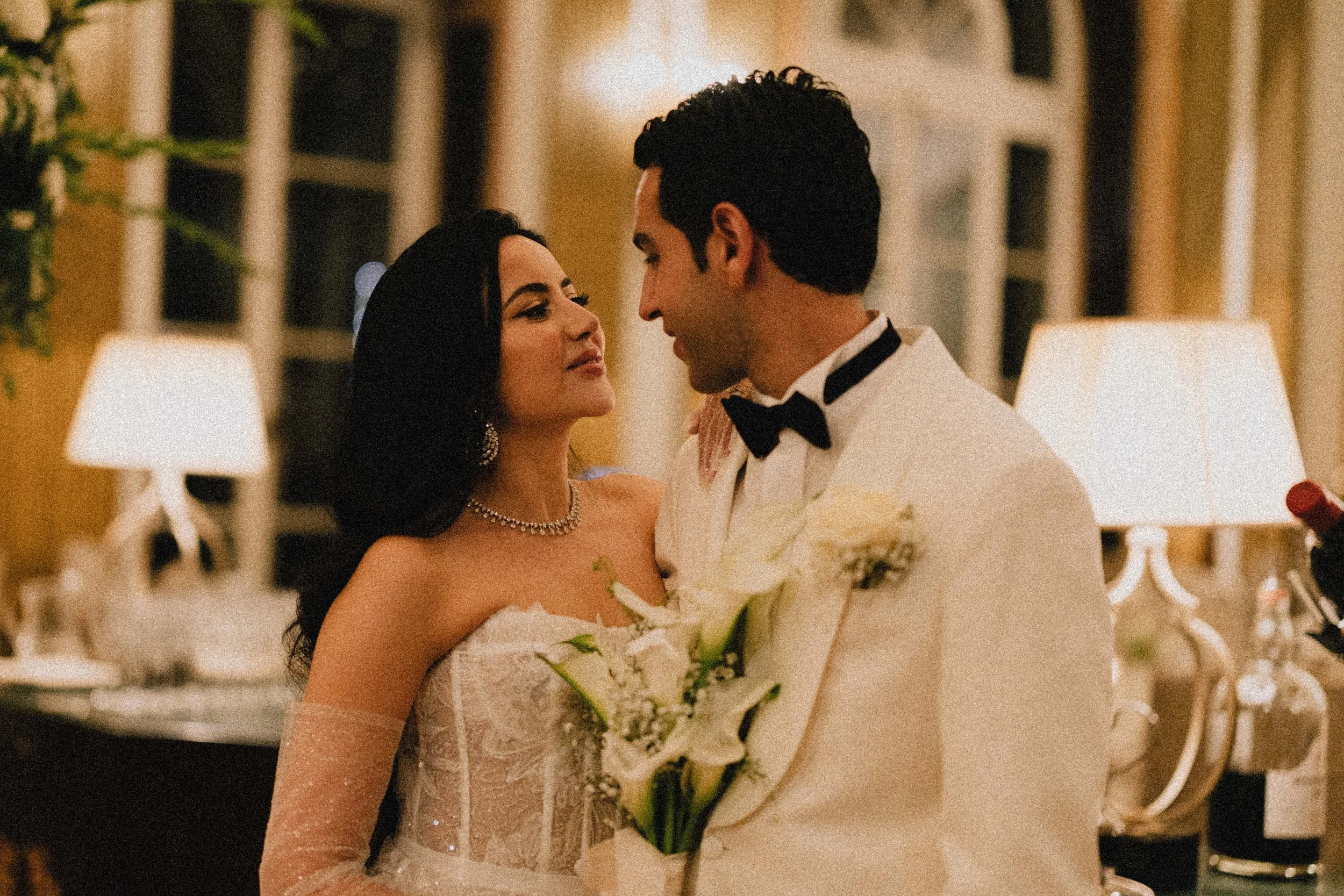 A newlywed couple sharing a romantic moment at their wedding reception, with the bride holding a bouquet of white calla lilies and both looking into each other's eyes in an elegant, warmly lit room.